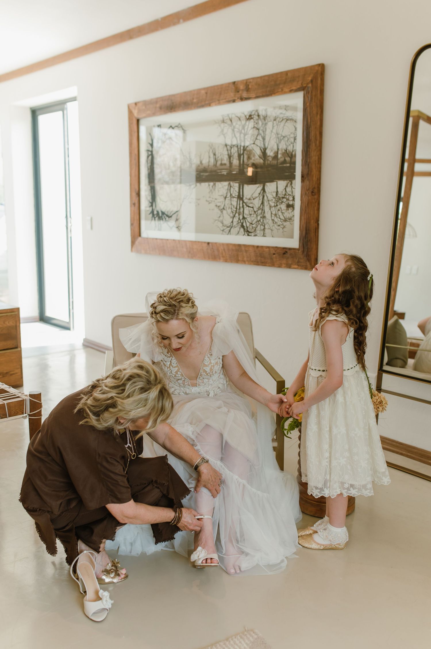 Wedding party members help with dress preparations in a sunlit room with wooden framed mirrors.