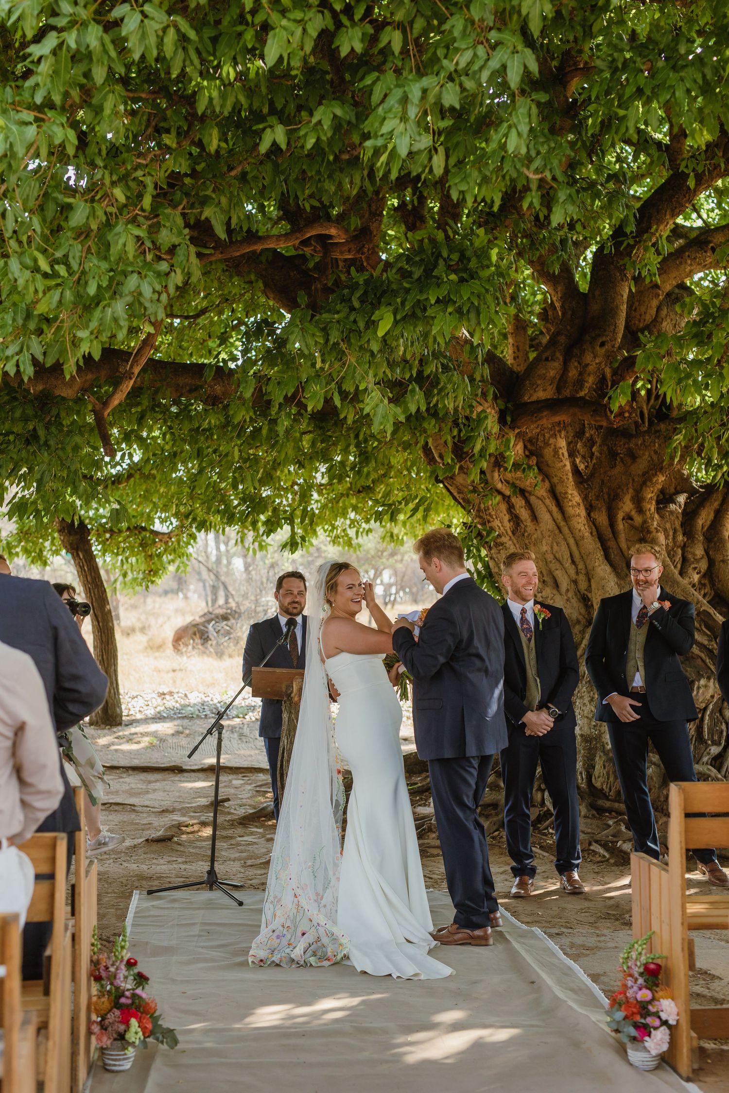 An intimate wedding ceremony unfolds beneath a majestic tree with wooden bench seating.