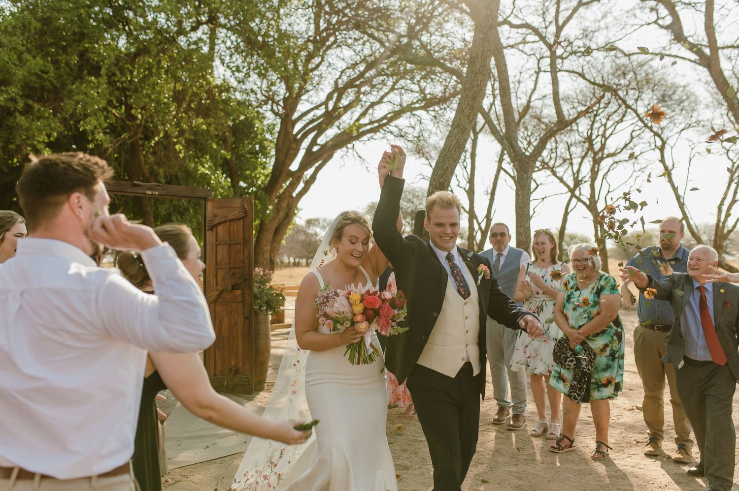 Wedding guests gather under trees at an outdoor ceremony during golden hour.