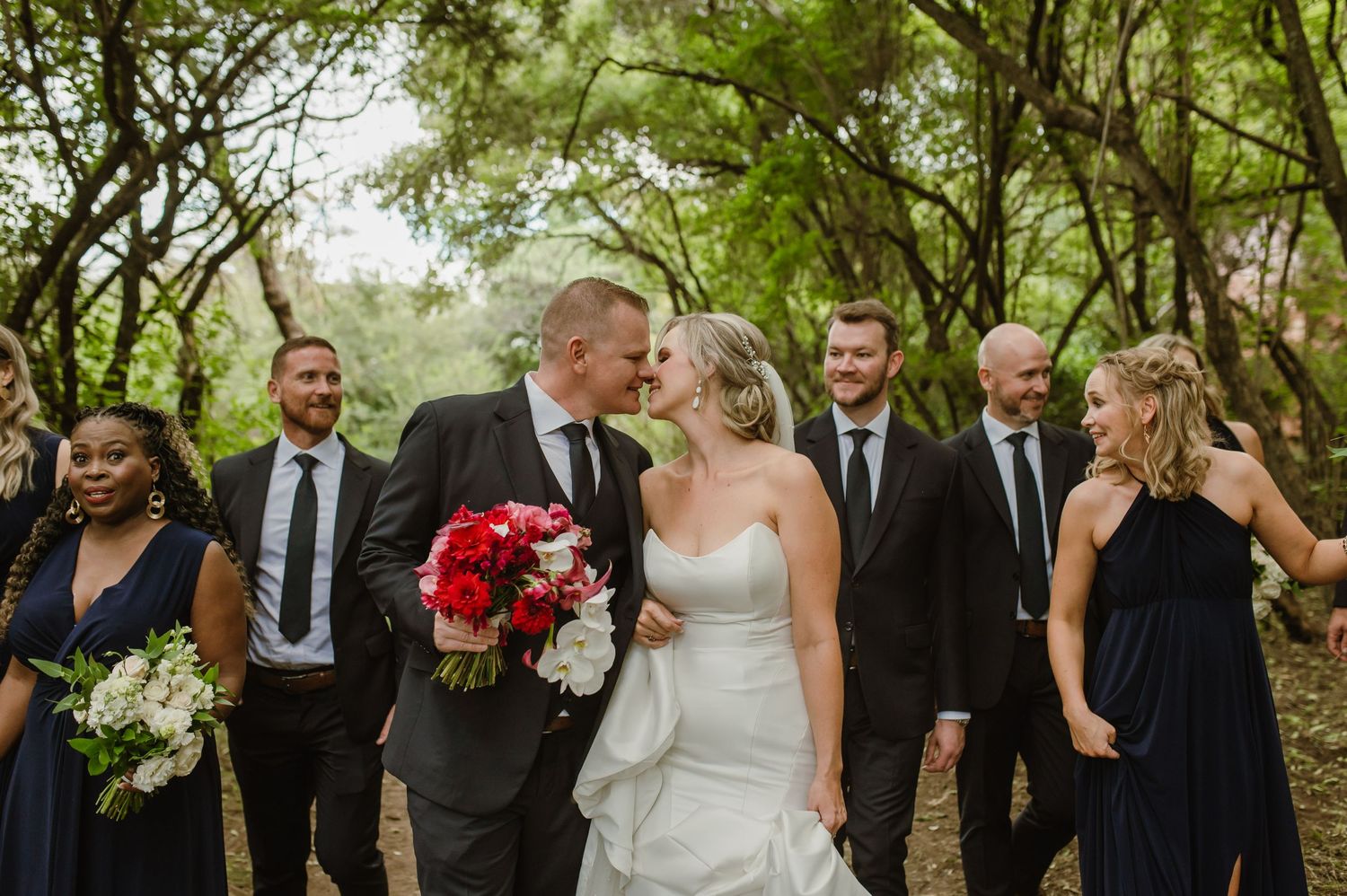 A wedding party shares a candid moment while walking through a tree-lined path.