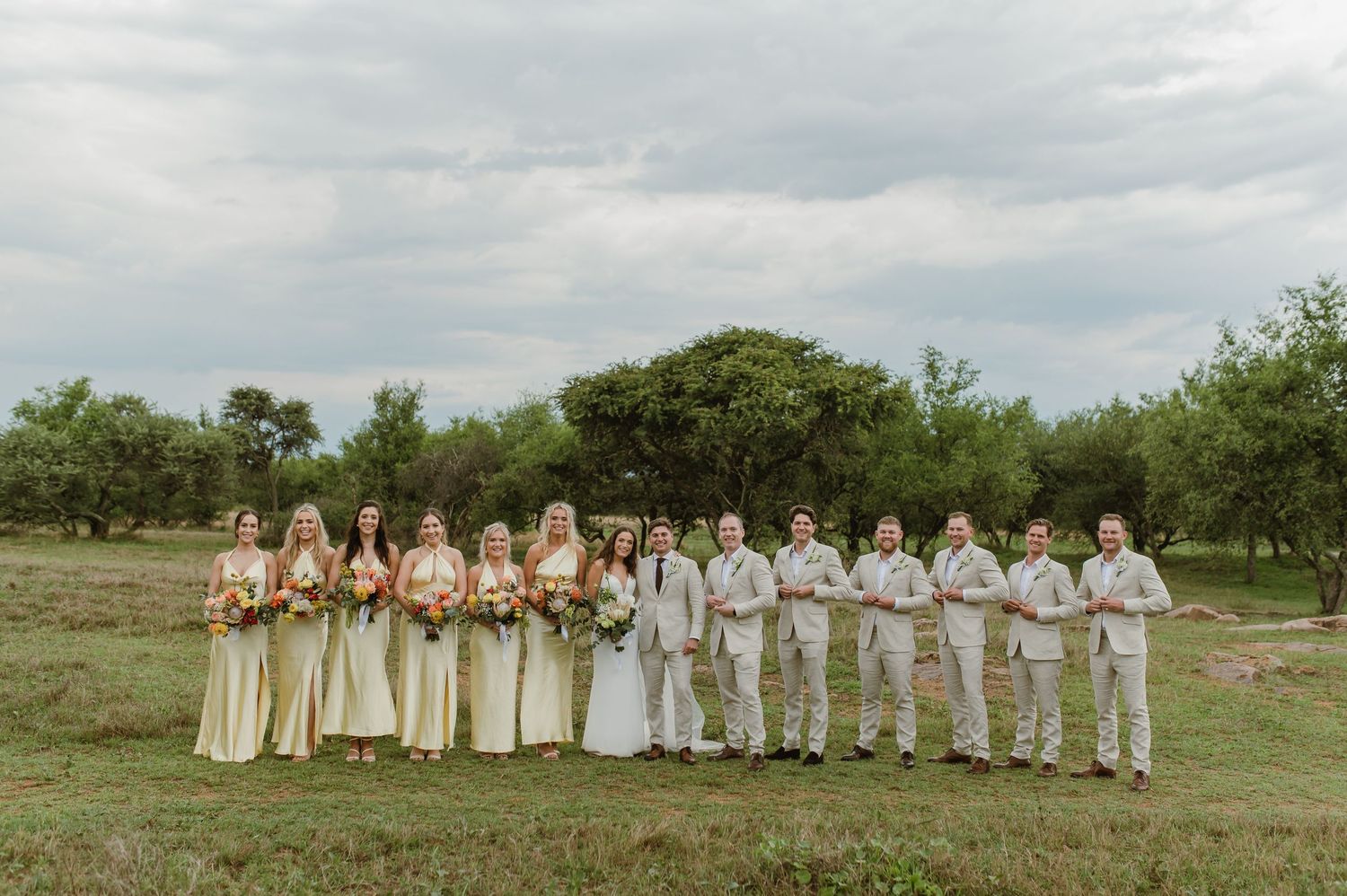 Wedding party in cream and white attire posed in a line against cloudy sky.