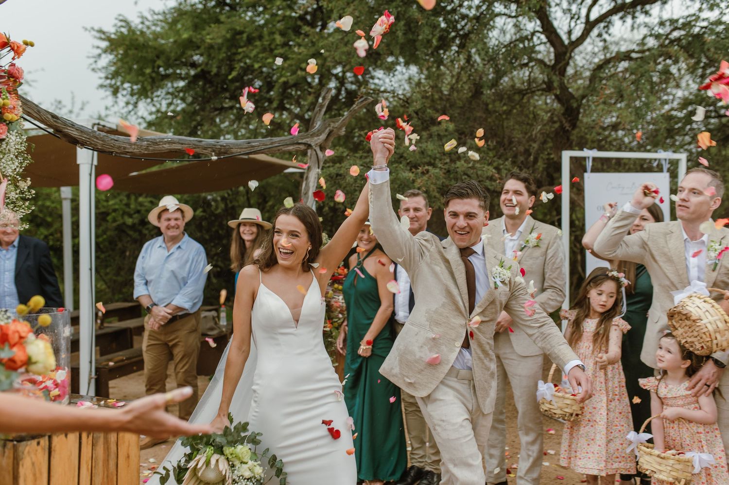 Wedding celebration with flower petals being tossed in the air under tree canopy.