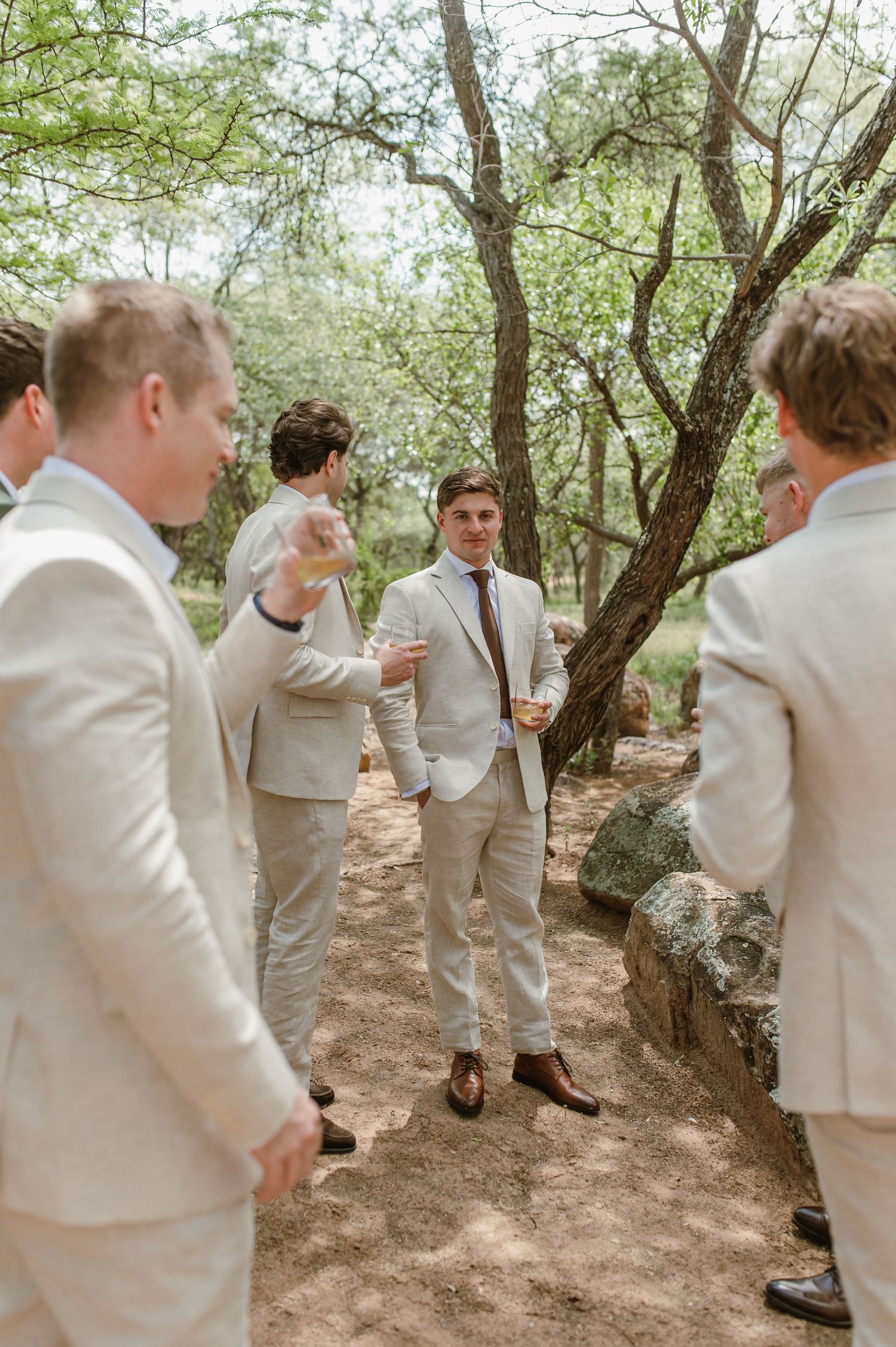 Group of groomsmen in light colored suits gather informally on a rustic outdoor path.