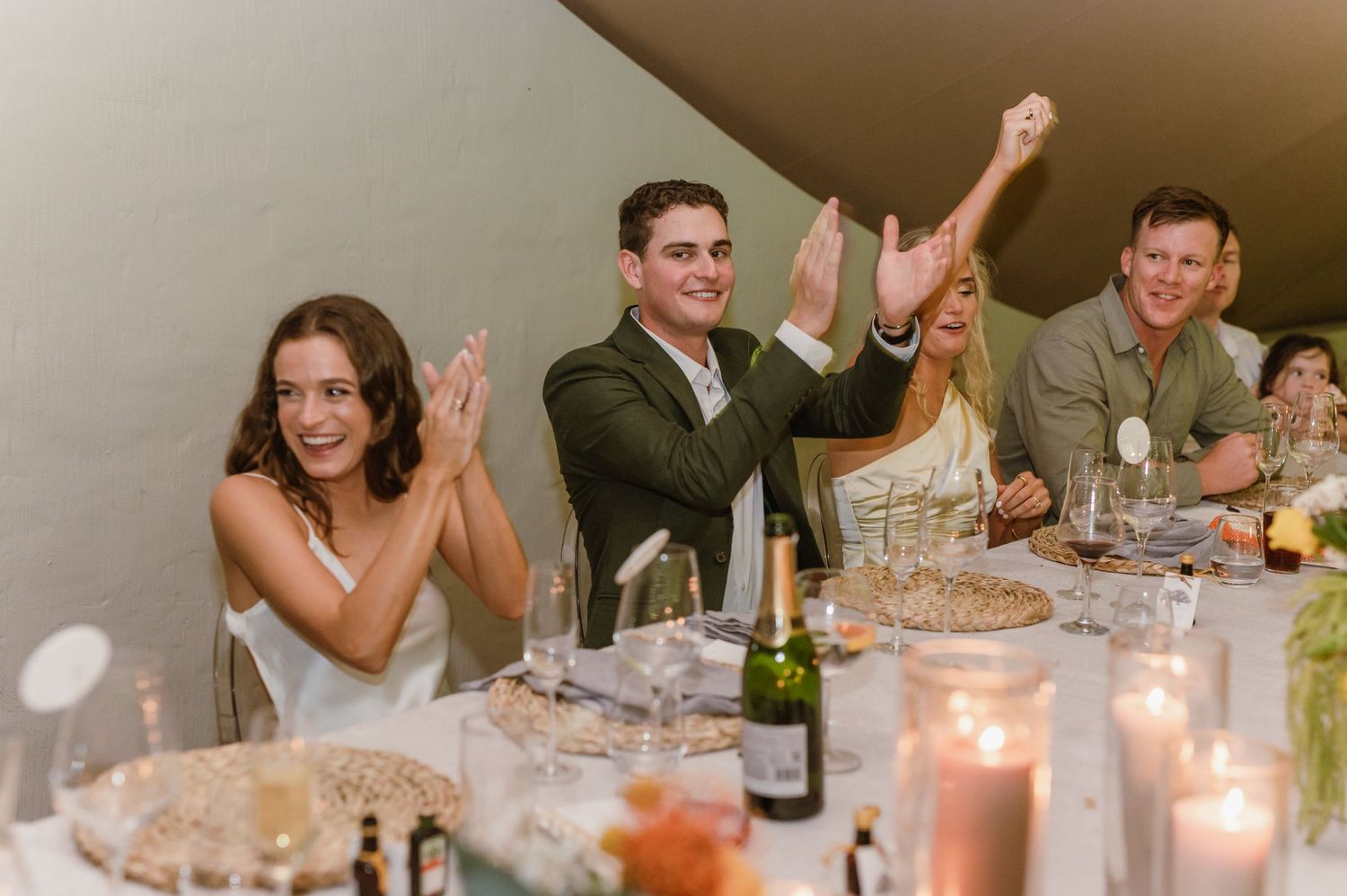 Wedding guests applauding and celebrating during reception toasts at candlelit dinner tables.