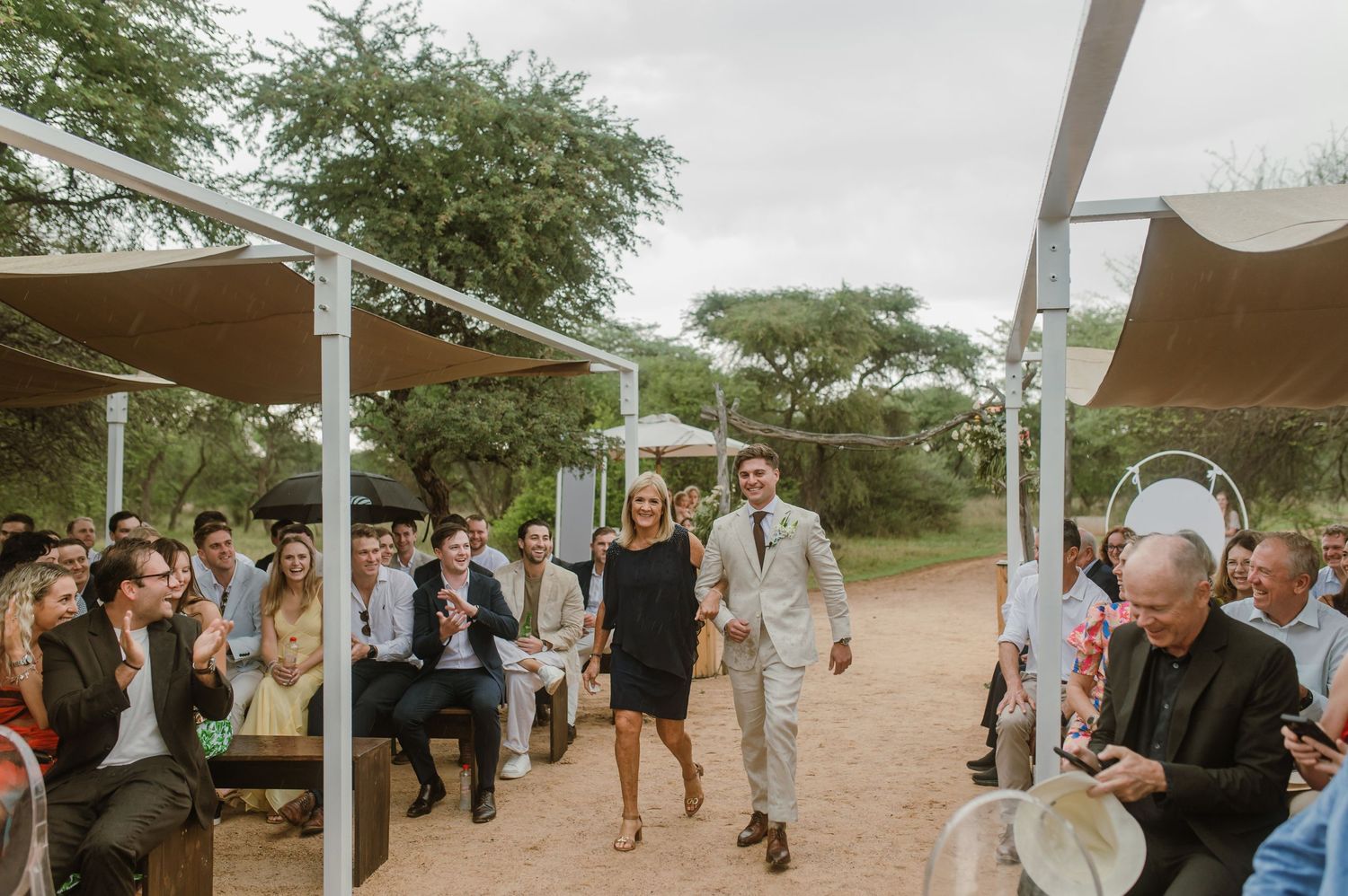 Wedding guests gather under trees at an outdoor desert venue with white canopy structures.