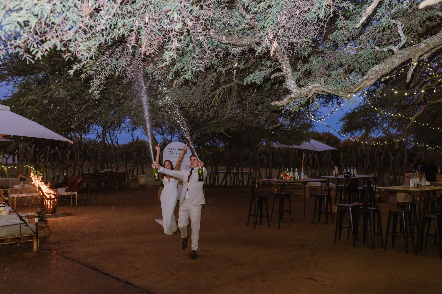 Empty chairs and tables set up under string lights for an outdoor wedding reception.