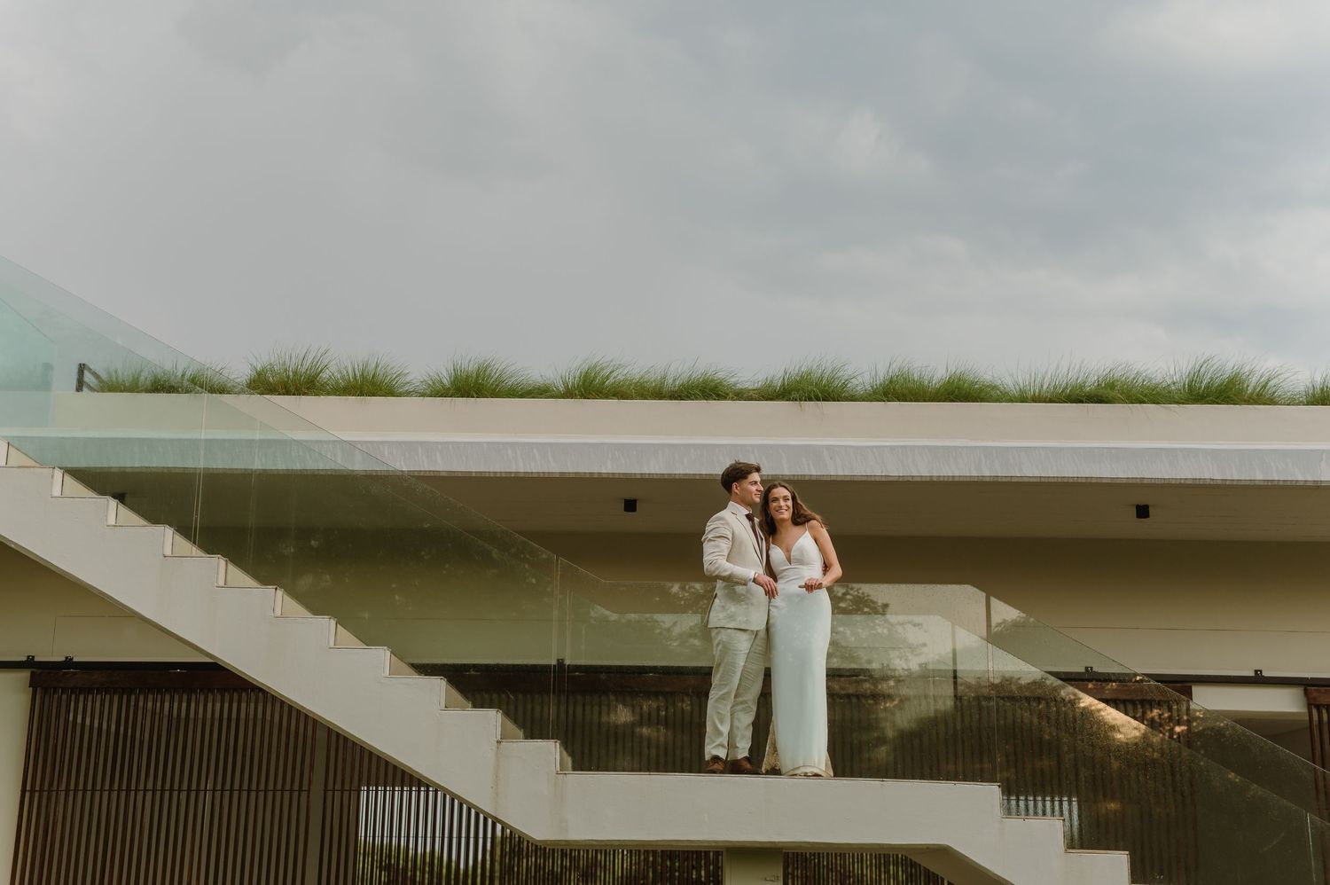 Wedding couple photographed on concrete steps of a modern building with green landscape backdrop.