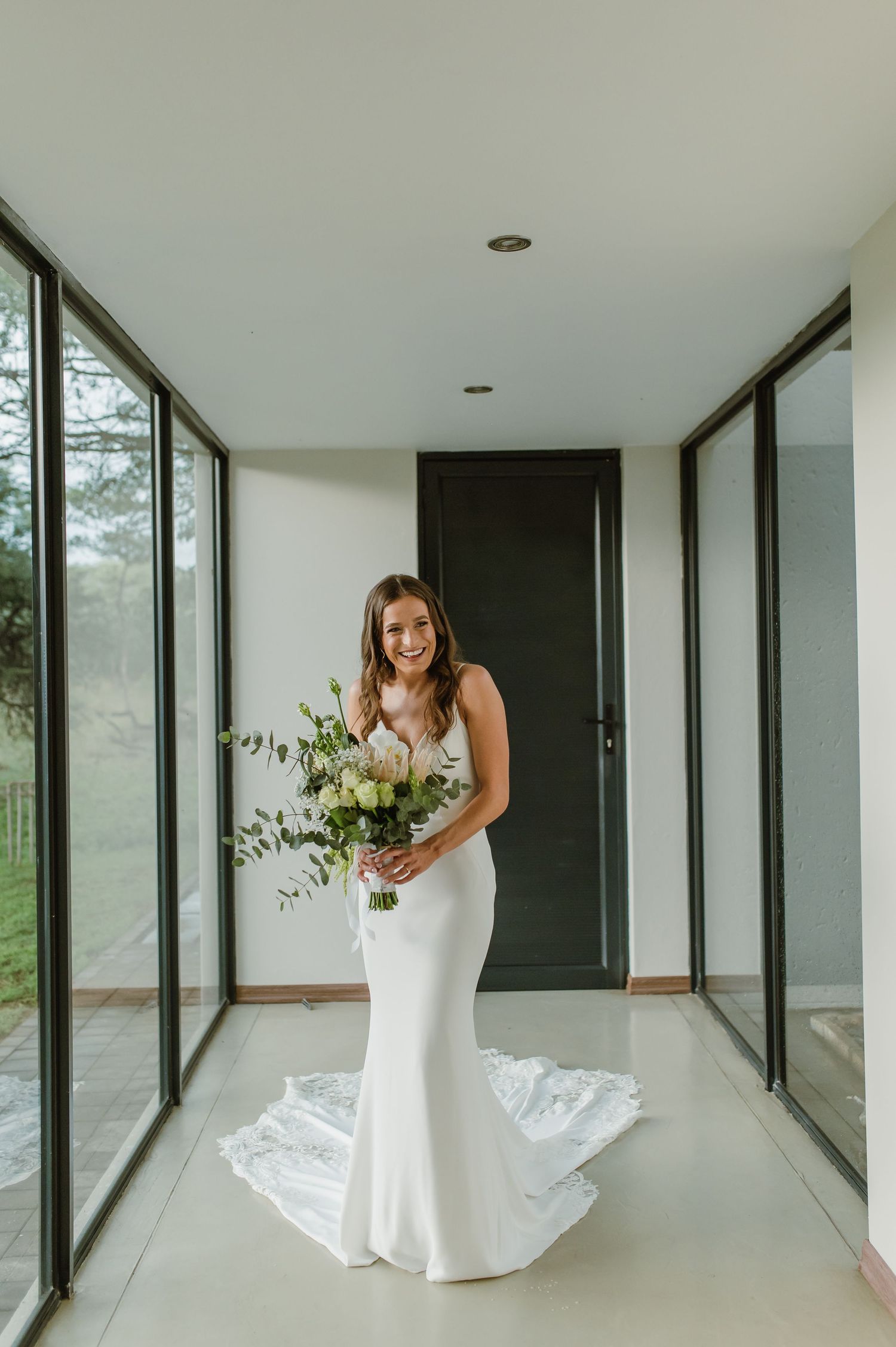 A person in a white wedding dress holds a bouquet while standing in a modern hallway with large windows.