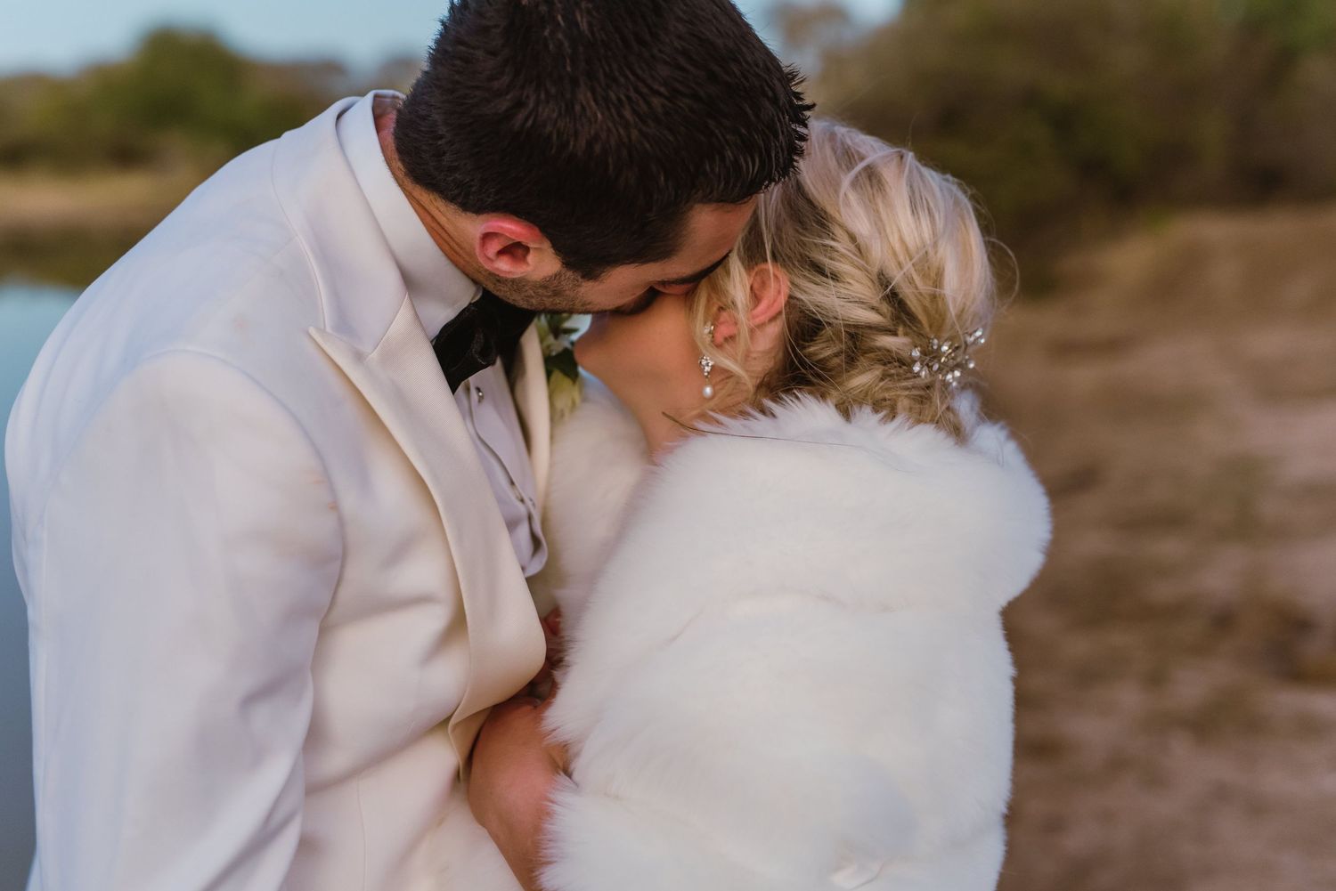 Intimate wedding couple embrace against a waterfront backdrop at sunset.