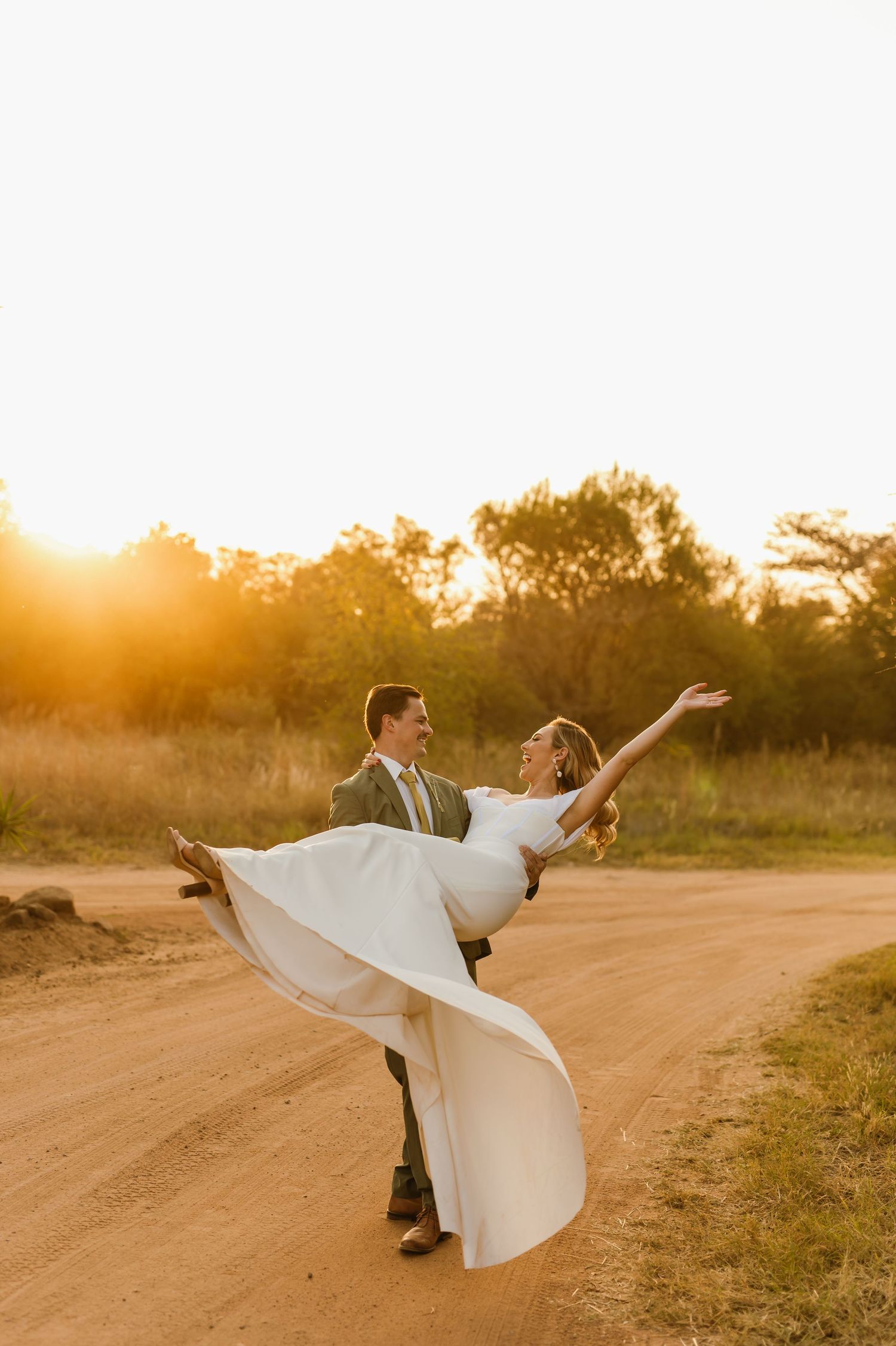 A couple spreads their arms in joyful celebration on a dirt road at sunset.