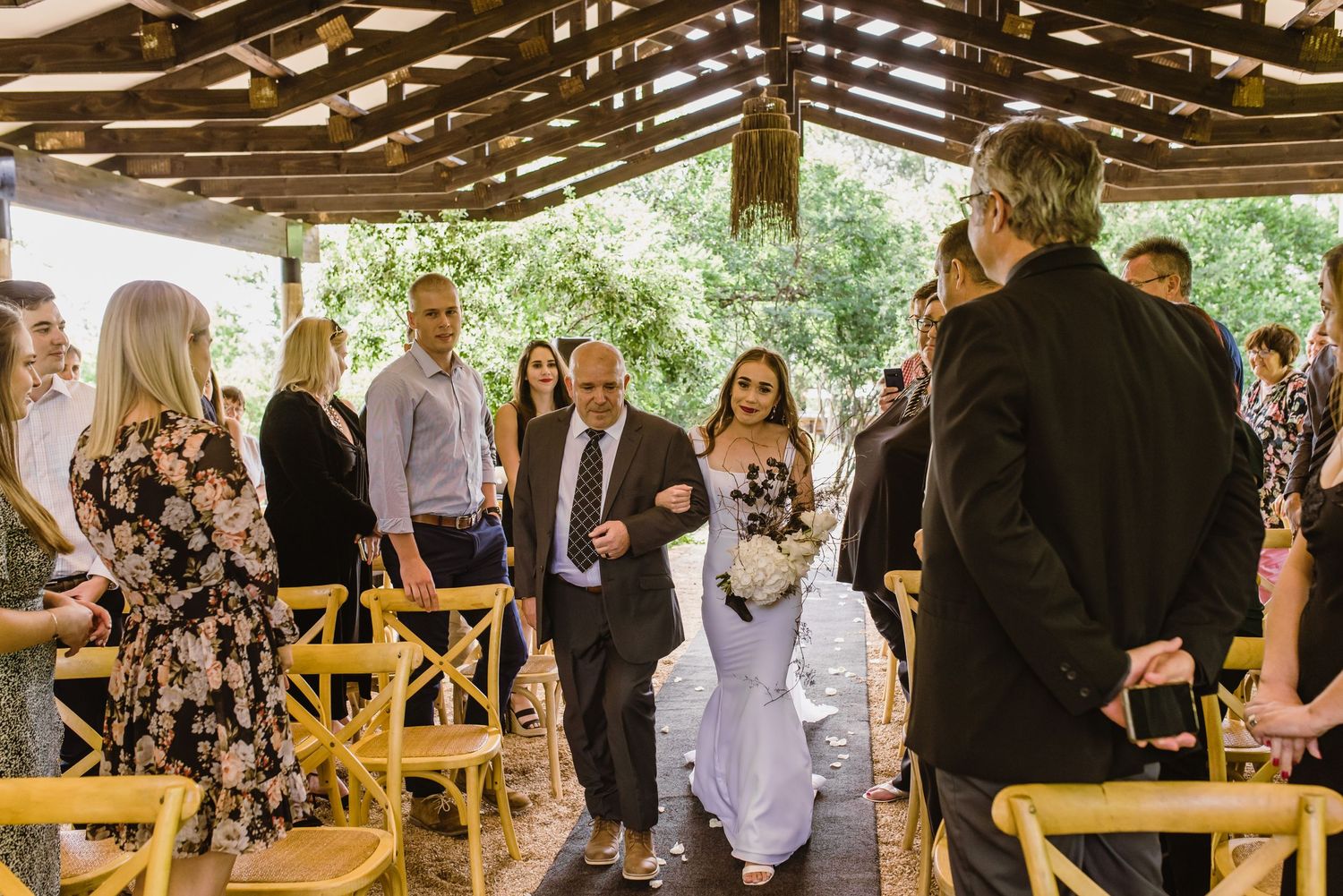 Wedding ceremony in progress under wooden pavilion with guests seated on rustic chairs.