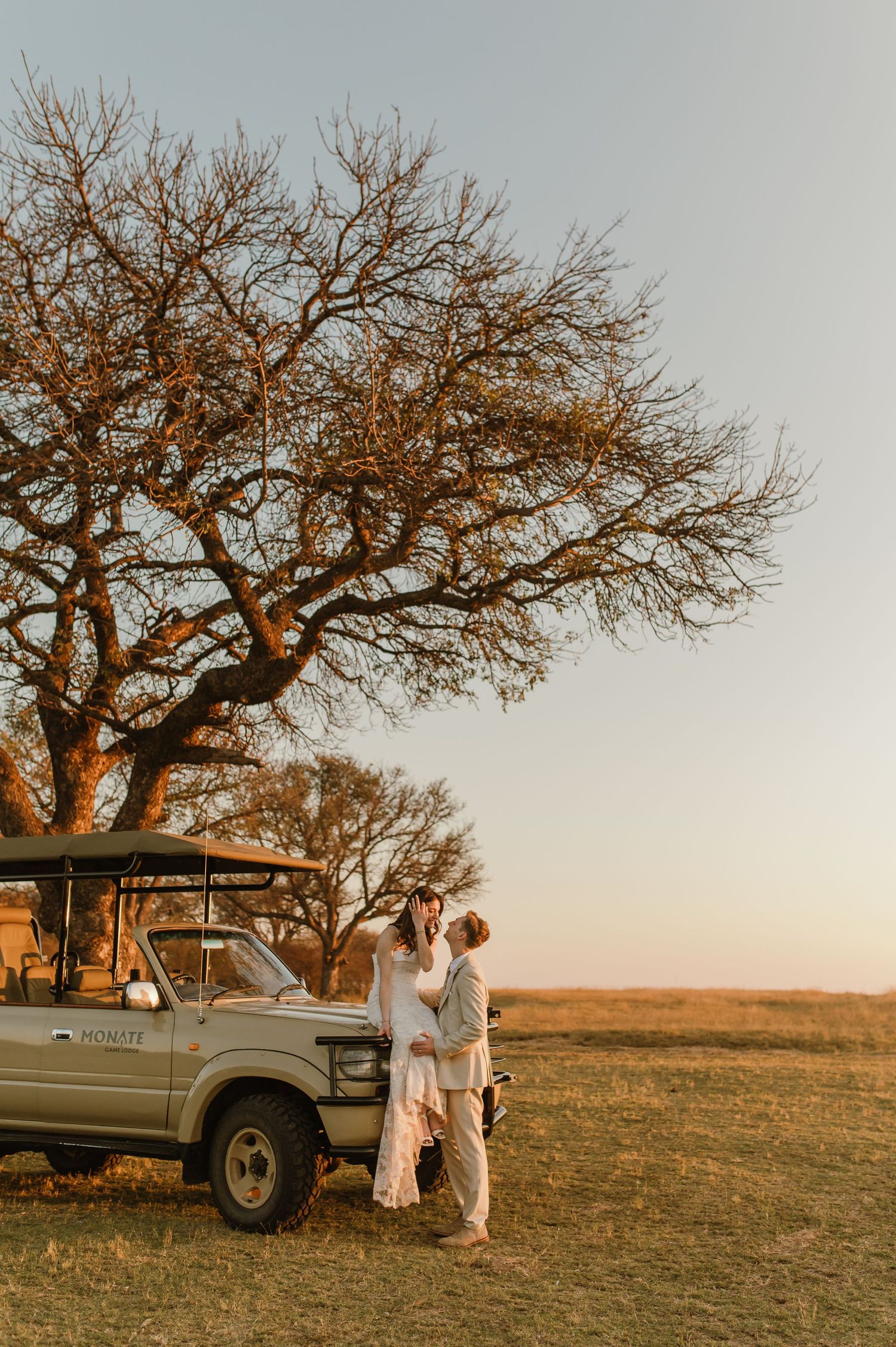 Couple embracing beside a safari jeep under a bare acacia tree at golden sunset in an African savanna.