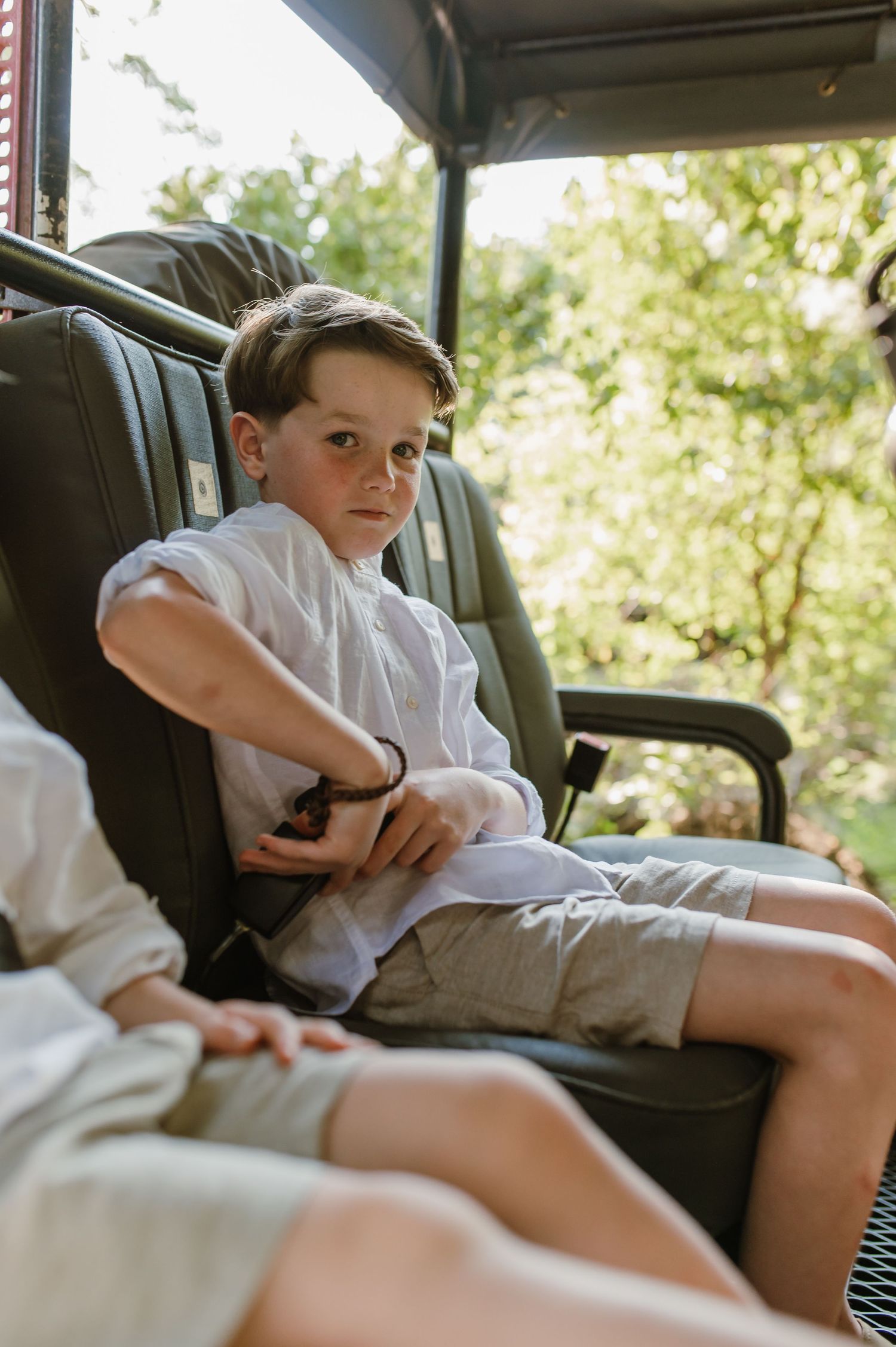 Young boy in white shirt seated in safari game drive vehicle looking at camera outdoors.