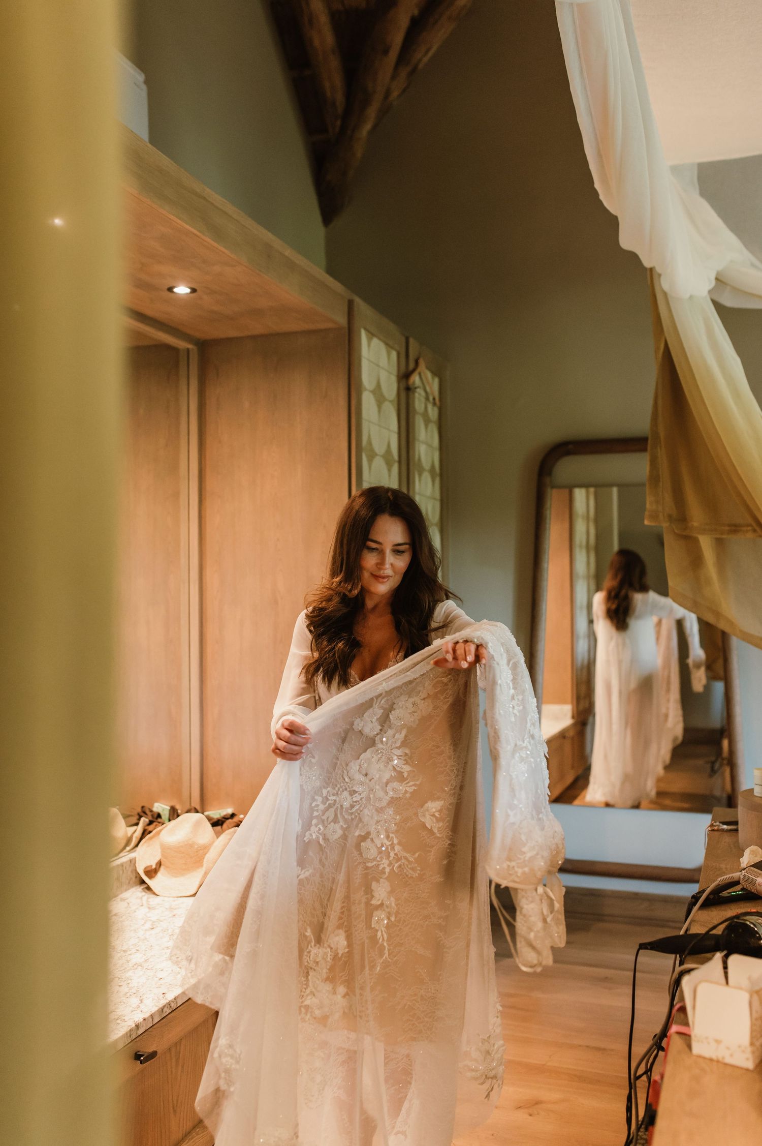 Bride in lace wedding gown holding her veil while getting ready in a warm-toned suite.
