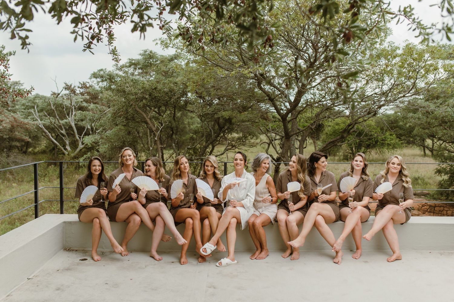 Wedding party sitting on concrete ledge outdoors with trees in background.