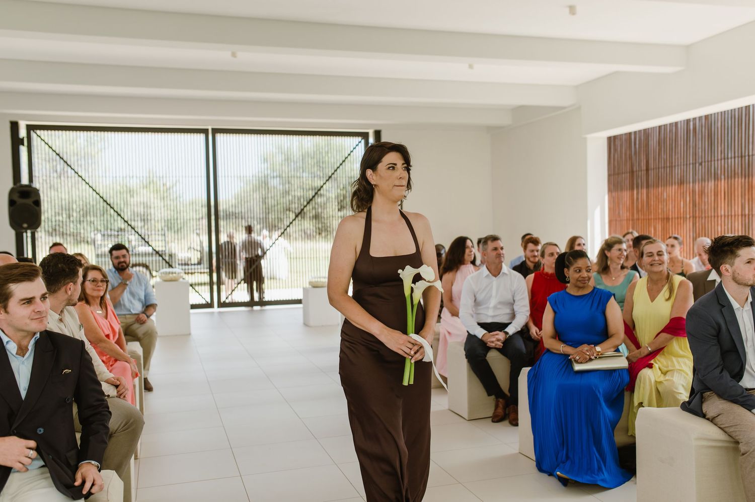 Wedding ceremony guests seated in modern venue with bridesmaid in brown dress walking down aisle.