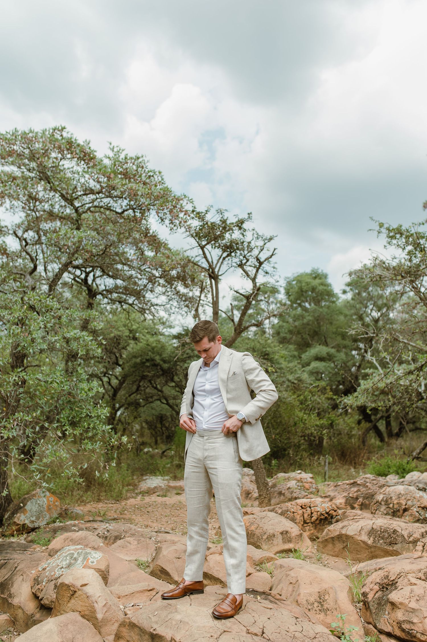 Person in light colored suit exploring rocky terrain under oak trees on cloudy day.