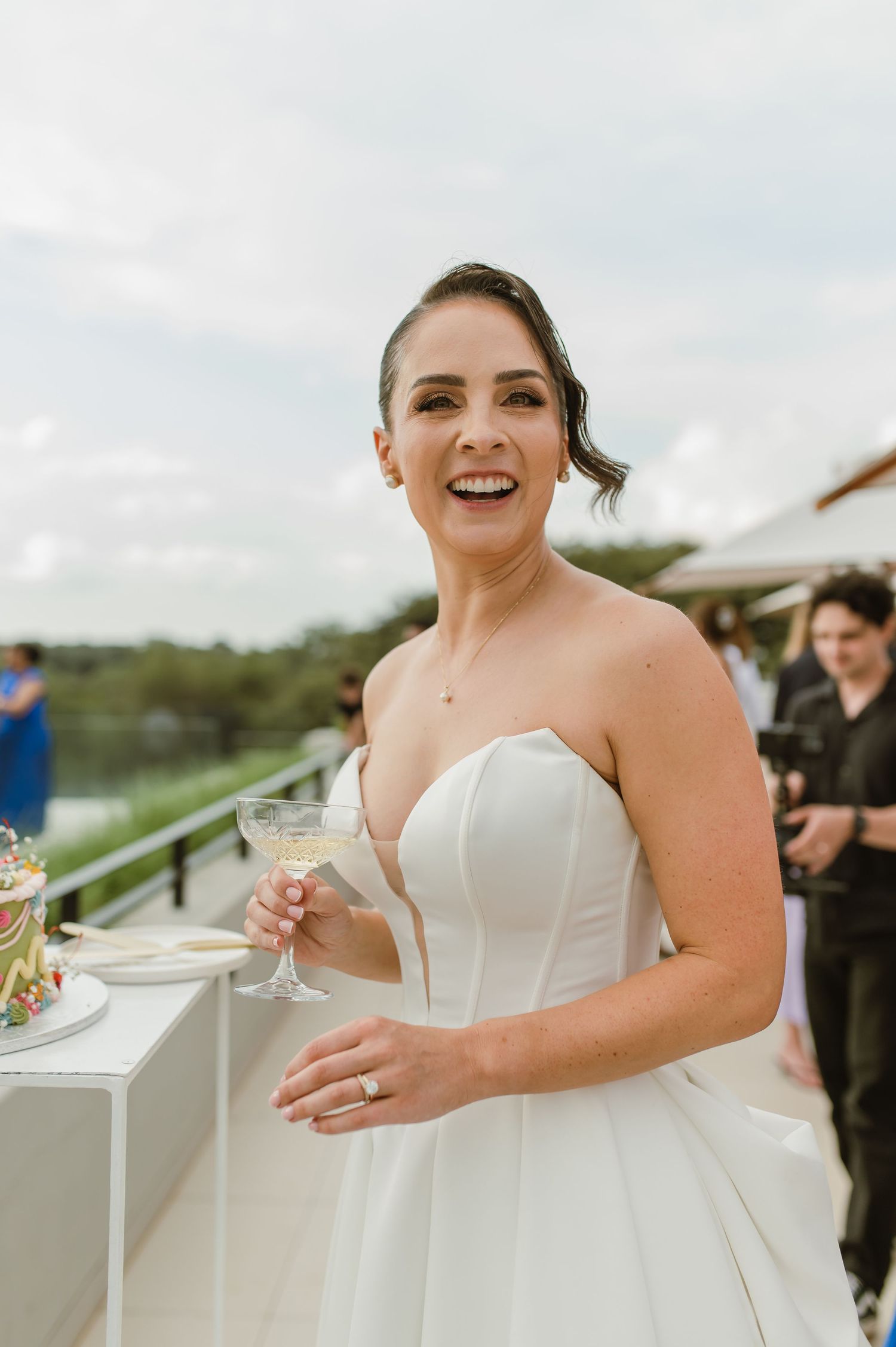 A person in a white strapless wedding gown holds a champagne glass during an outdoor celebration.