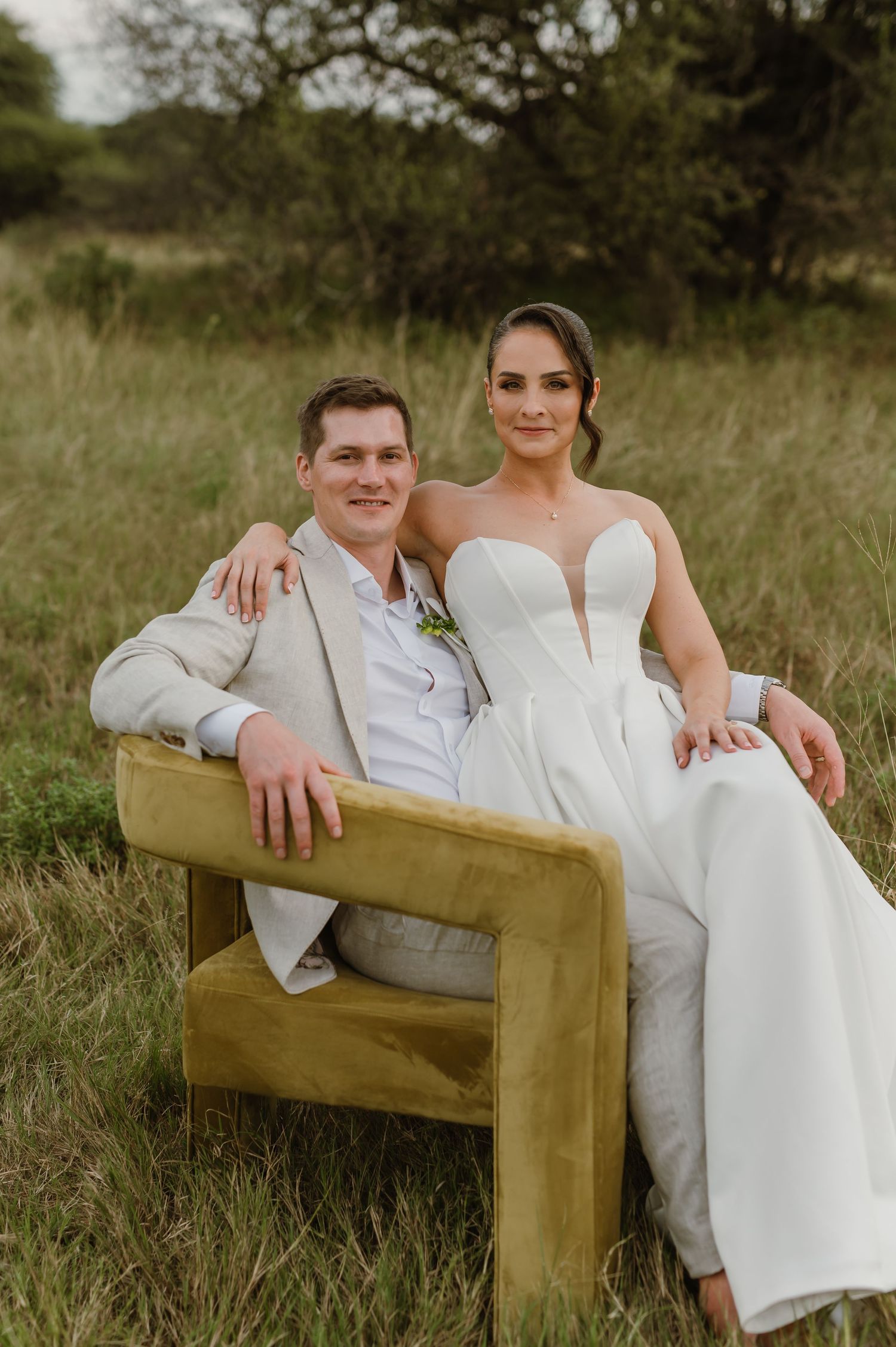 A couple sits together on a rustic wooden bench in a grassy field during a wedding photoshoot.