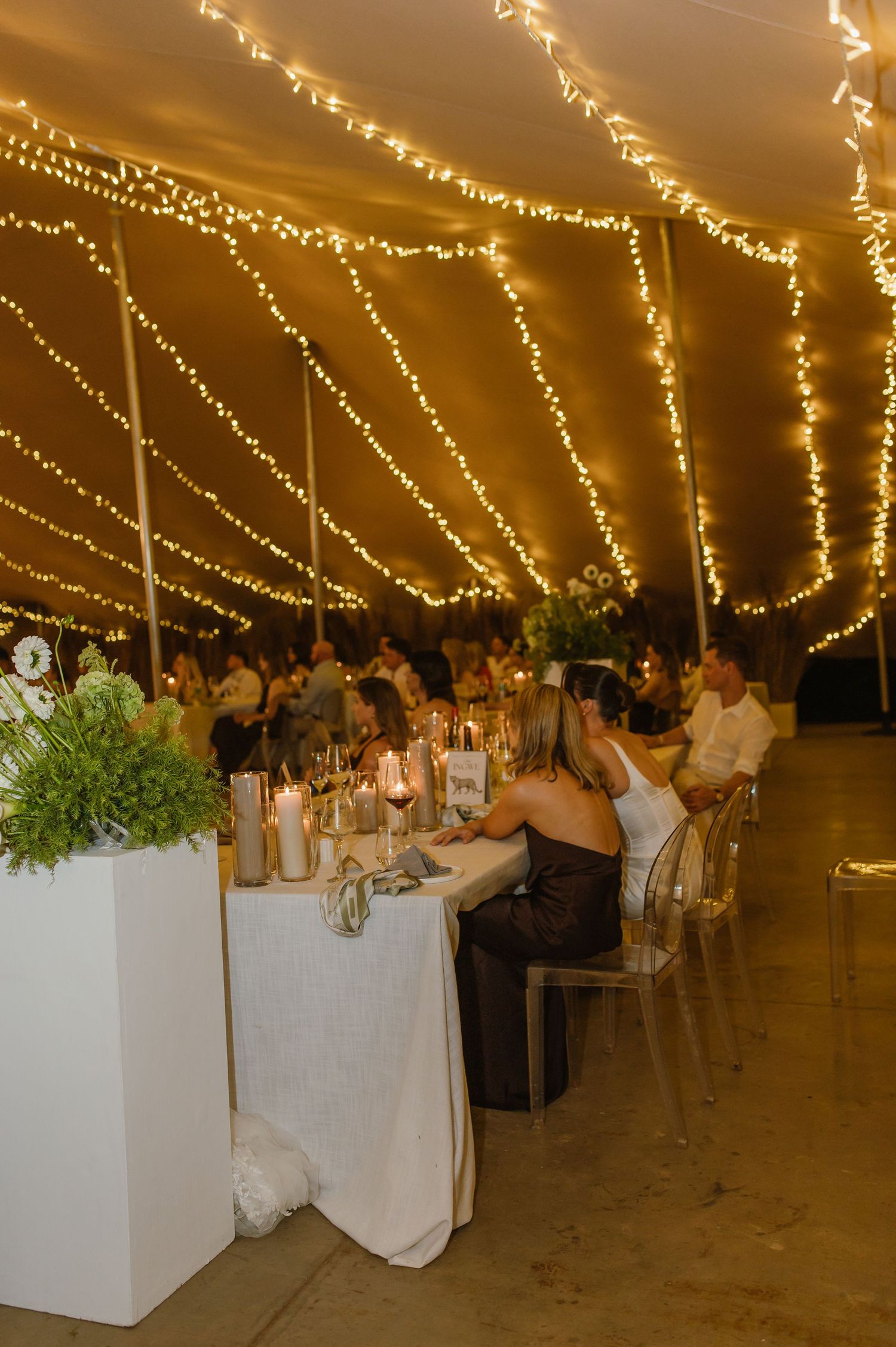 Wedding reception tent decorated with string lights and white floral arrangements.
