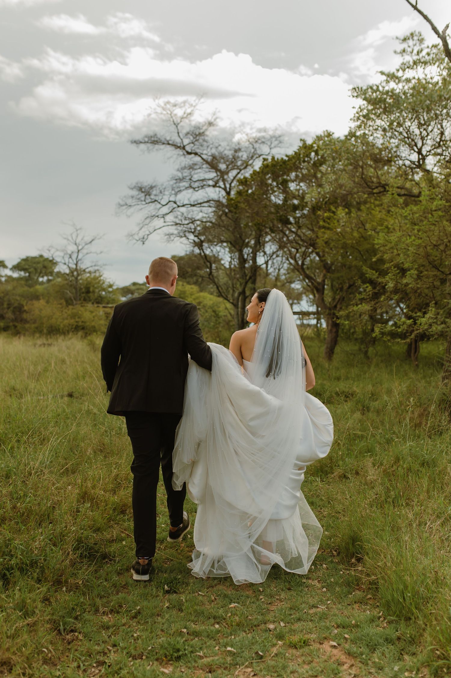 Bride and groom walk hand in hand through green meadow, wedding veil flowing in the breeze.