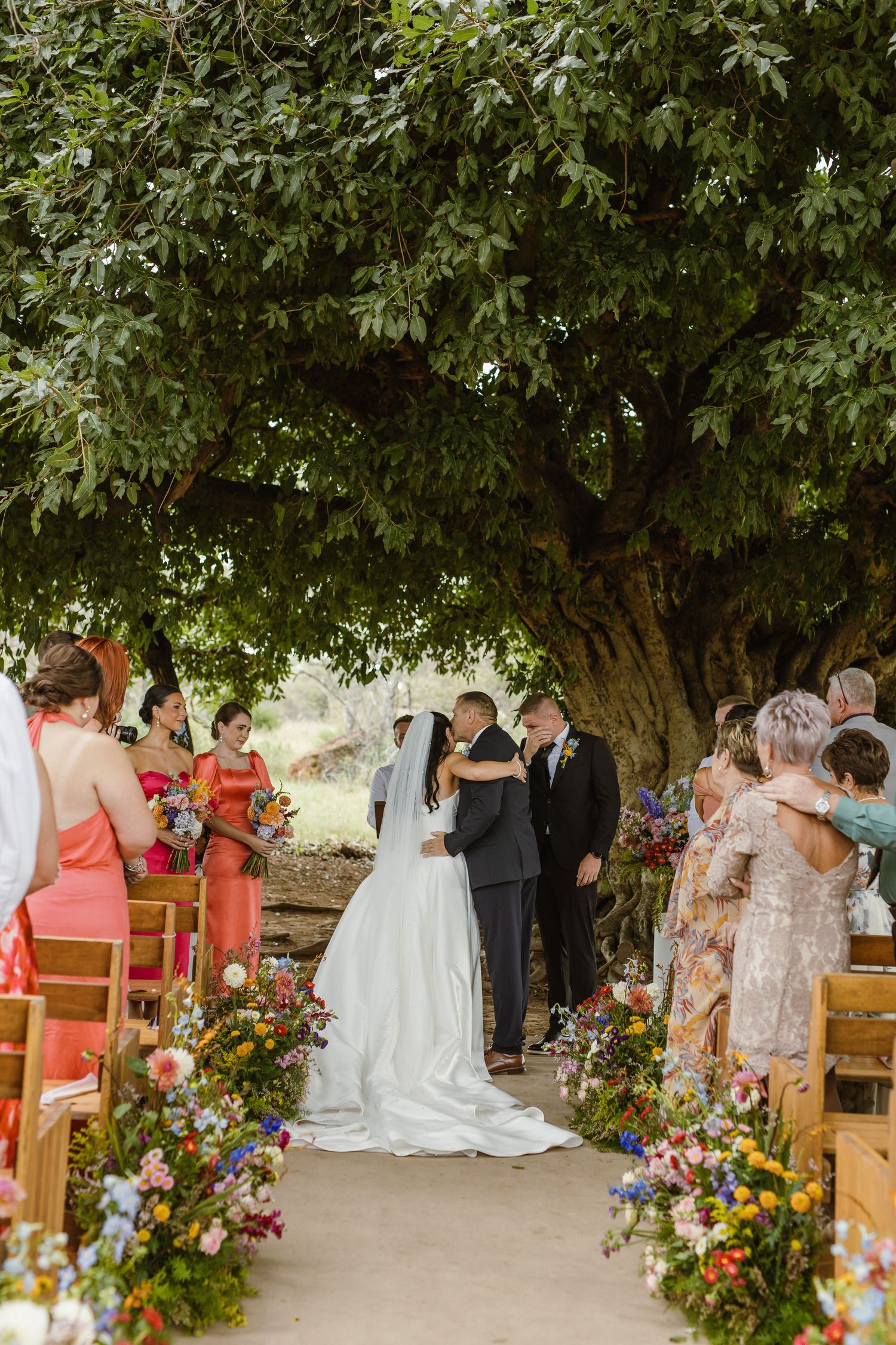 Outdoor wedding ceremony under a large tree with colorful floral aisle decorations.