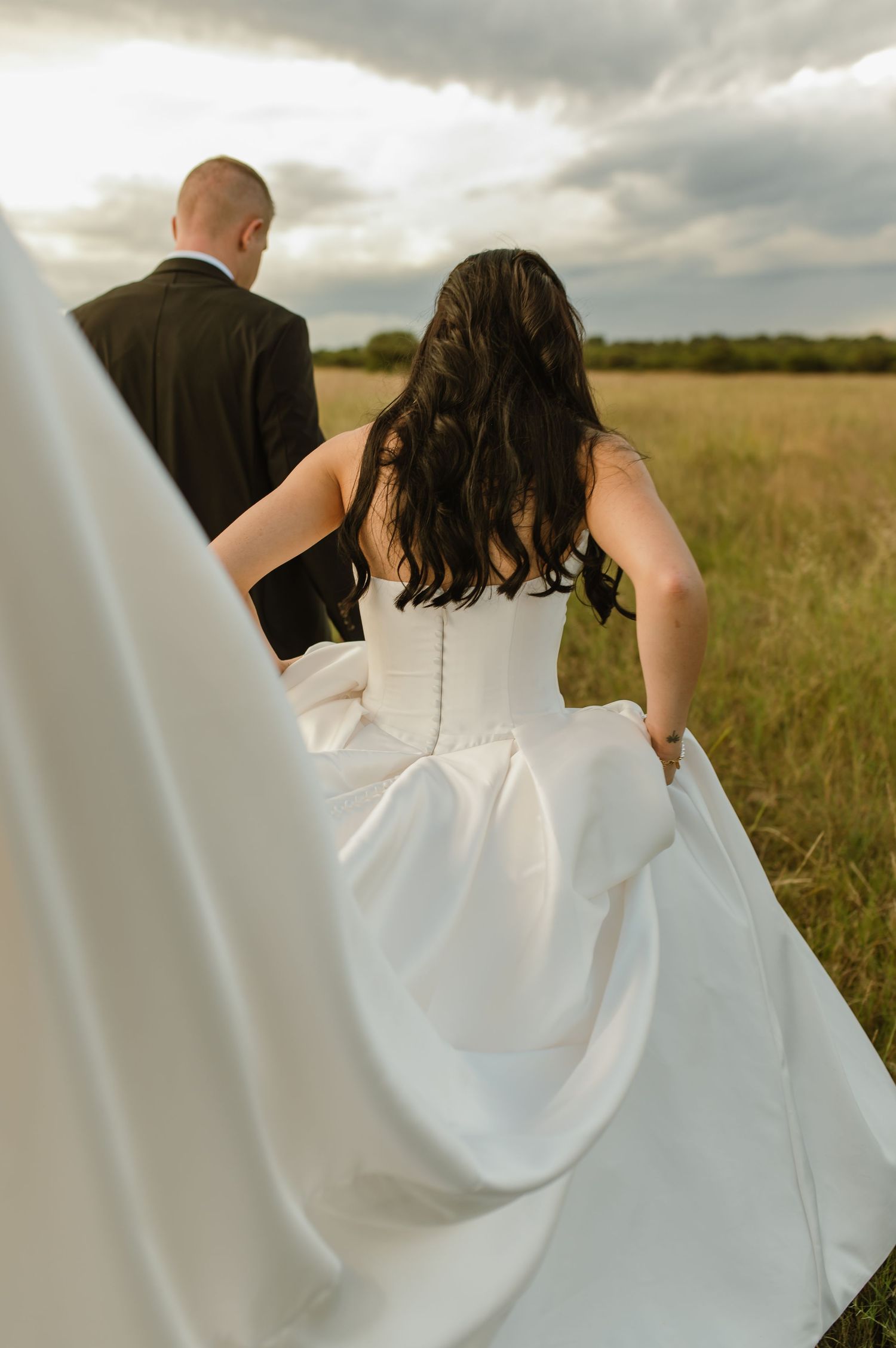 Bride in white satin gown and groom in black suit walk through open green field.
