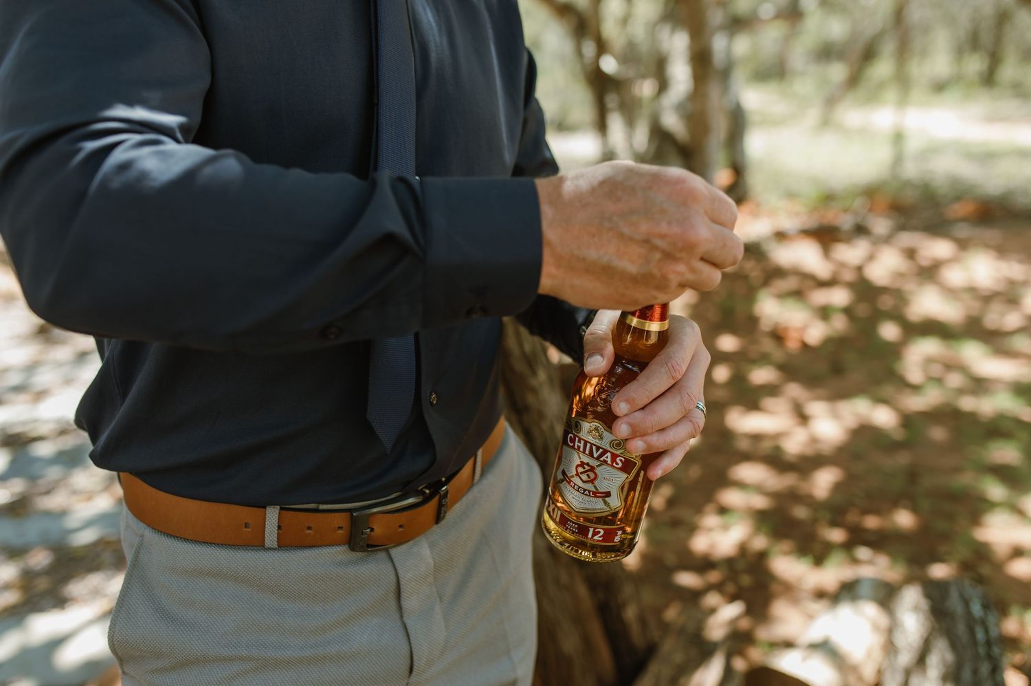 Man in dark shirt opening whiskey bottle outdoors among trees wearing suspenders.