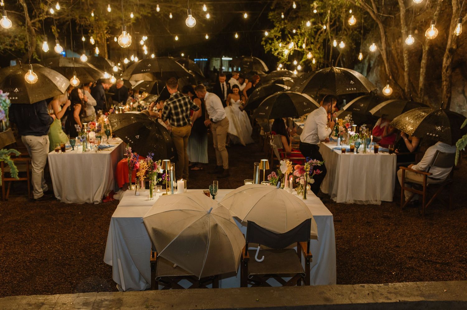 Elegant outdoor wedding reception at night with string lights and umbrella table decorations.