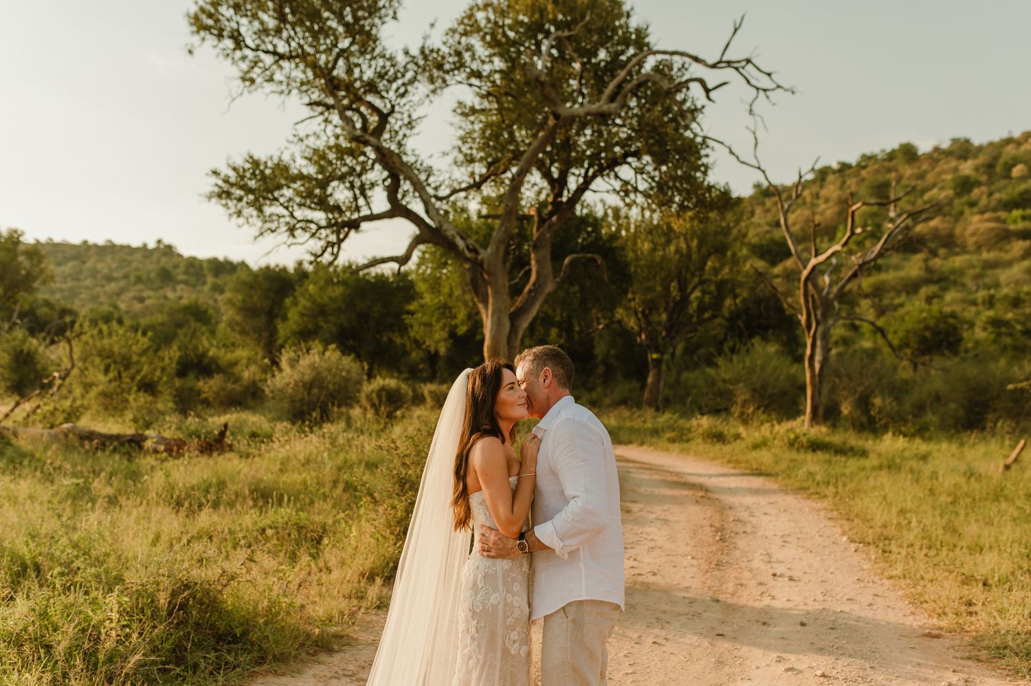 Bride and groom share a romantic kiss on a dirt path surrounded by lush greenery and trees during golden hour.