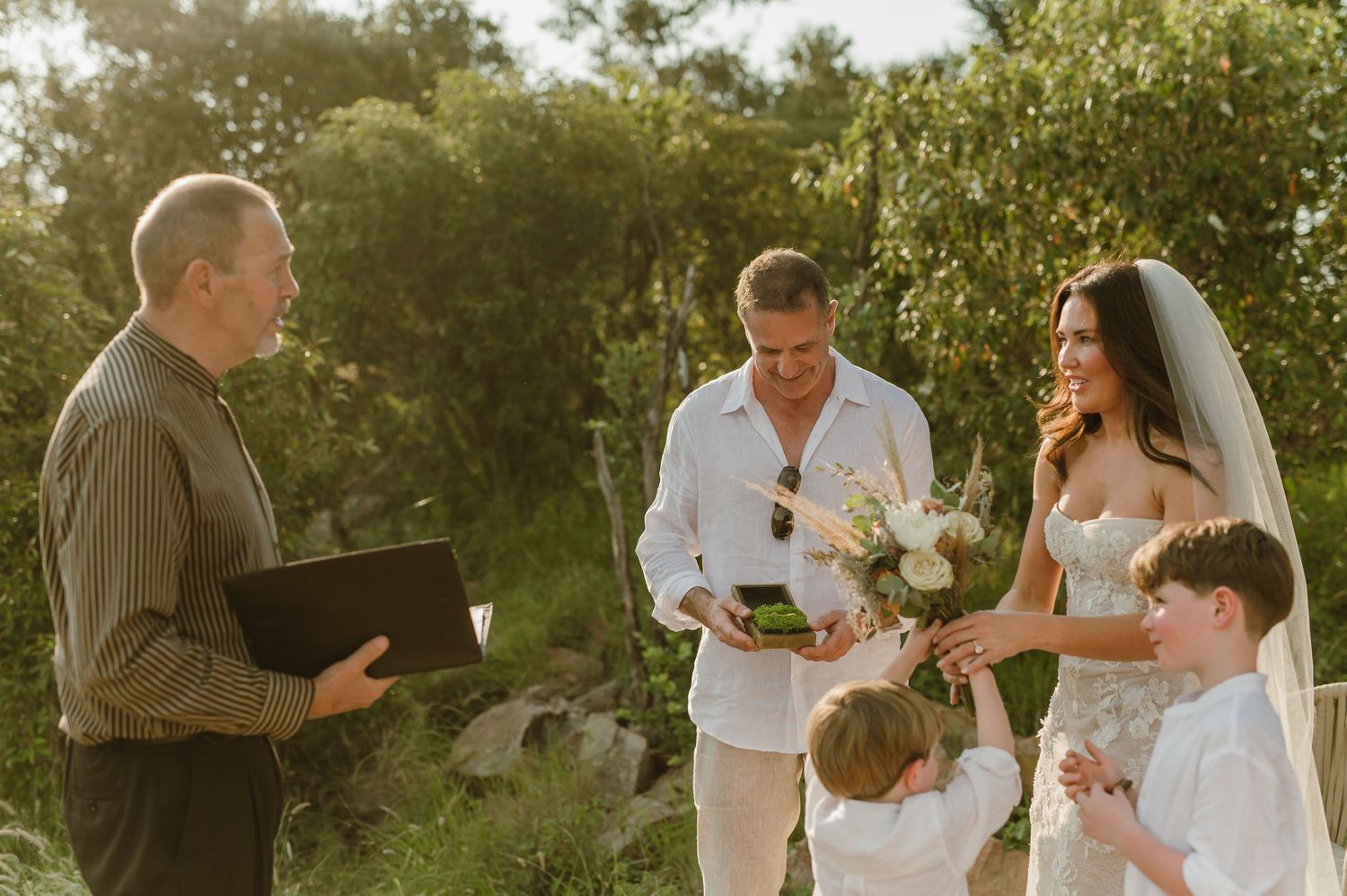 Outdoor wedding ceremony with officiant, groom in white, bride in veil holding bouquet, and two children in natural setting.