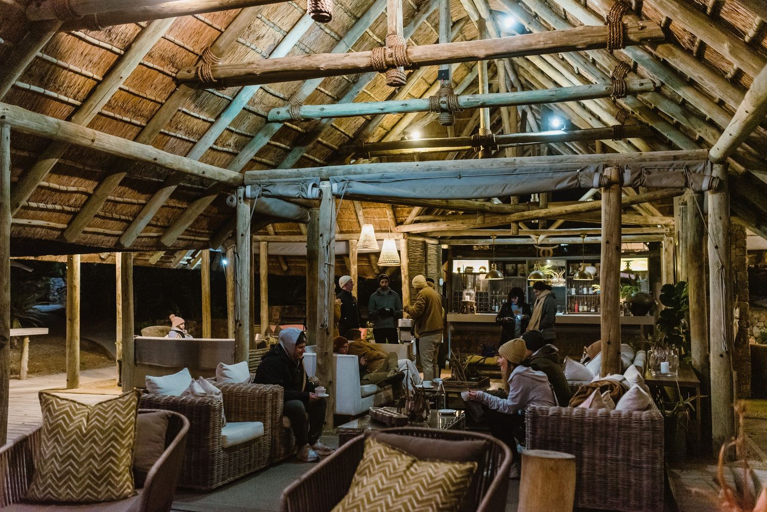 Cozy outdoor lounge area with thatched roof and comfortable wicker furniture illuminated by warm lighting at dusk.