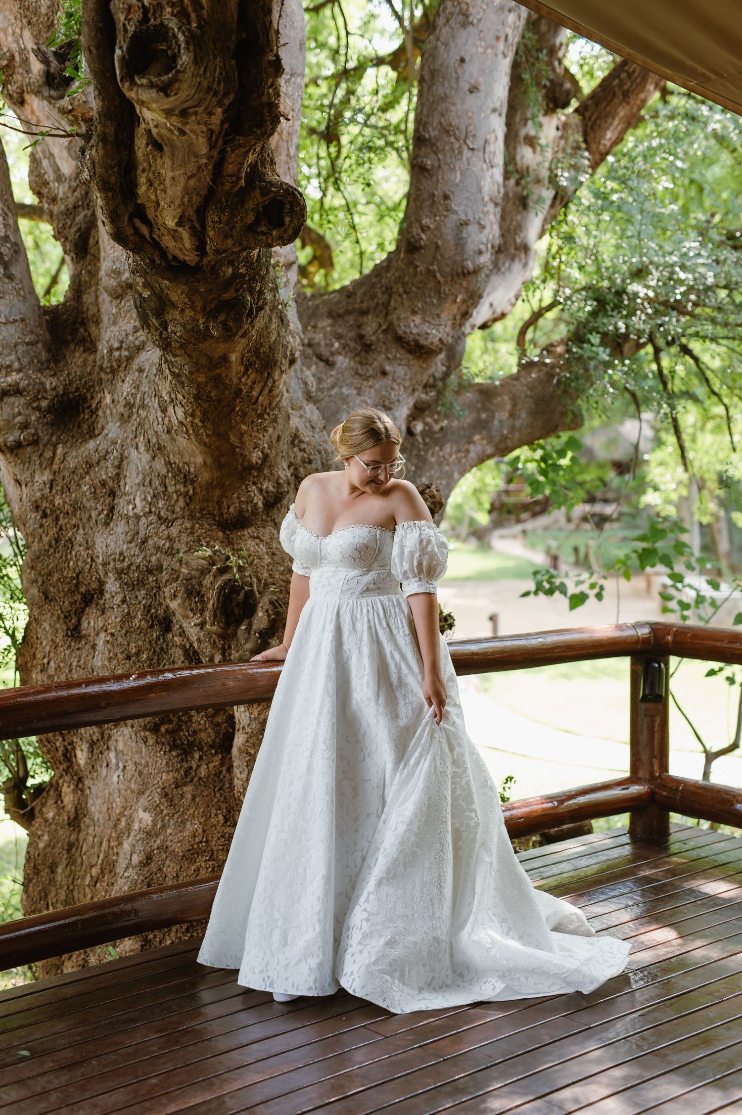 A flowing white wedding gown with puff sleeves and lace details is showcased on a wooden deck beneath a large tree.