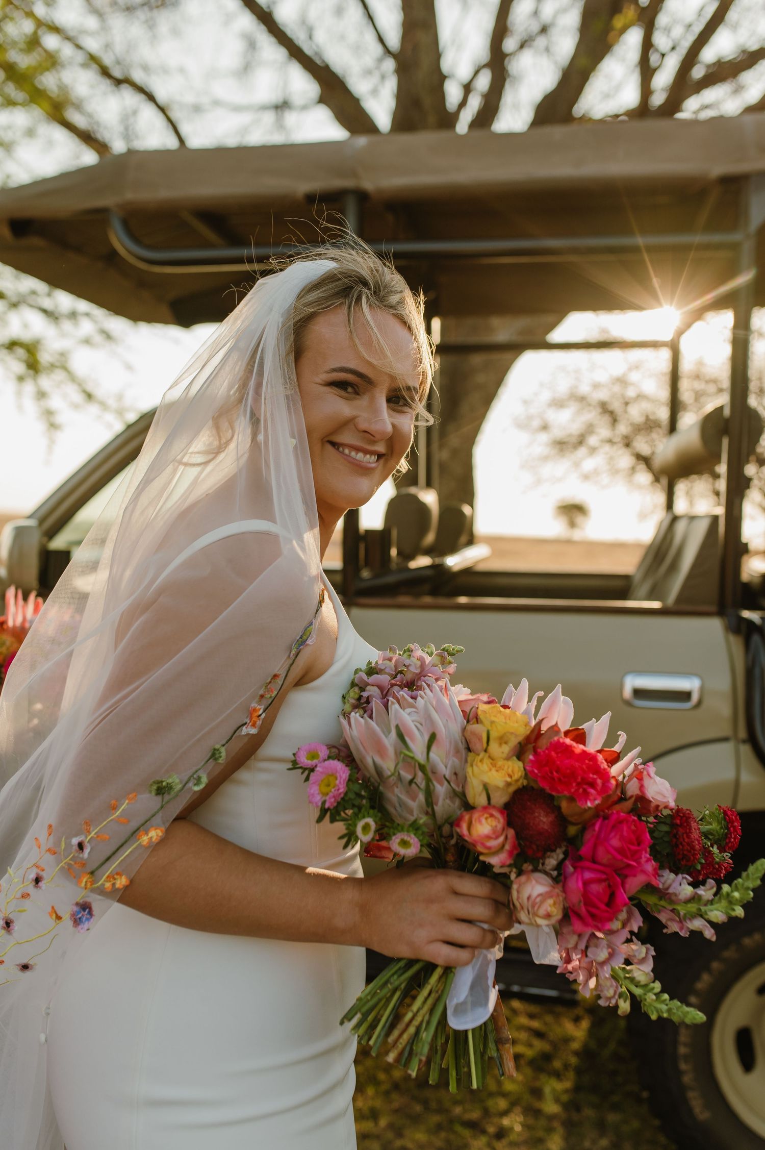 A bride in white dress holds a vibrant pink and red bouquet near a vintage vehicle.