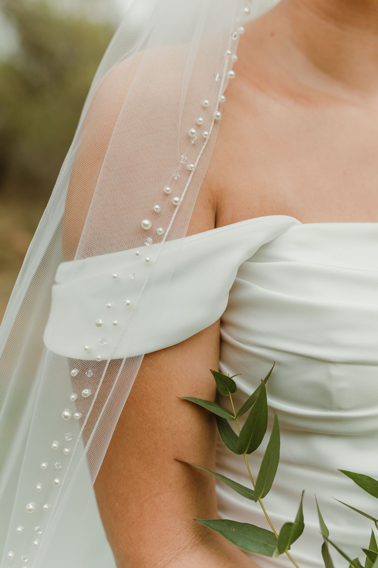 Close-up detail of pearl-adorned wedding veil and off-shoulder white bridal gown with greenery accent.