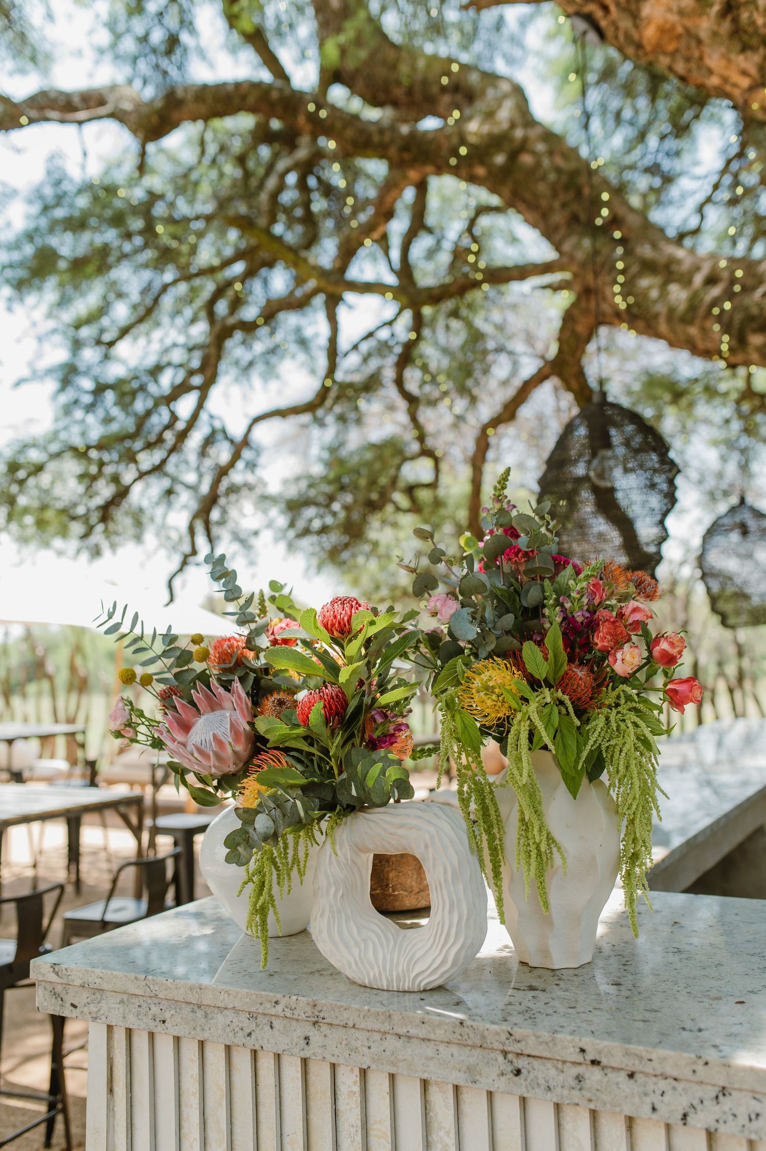 Colorful floral arrangements in white vases displayed on a vintage table under tree branches.