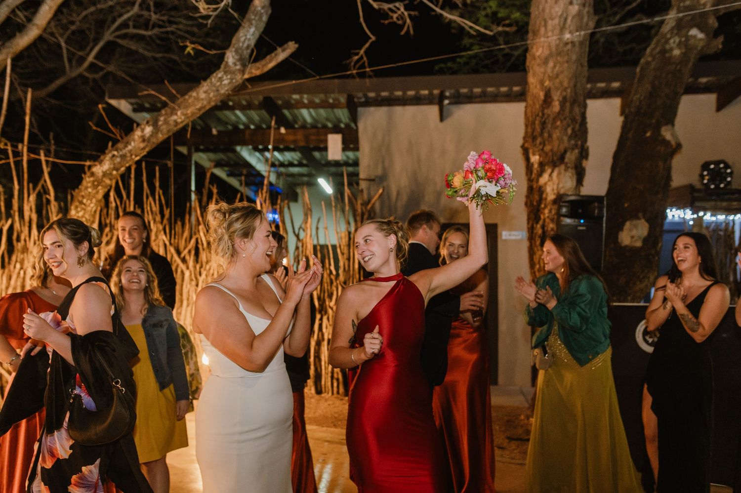Wedding party dancing and celebrating in a rustic outdoor venue illuminated by string lights.
