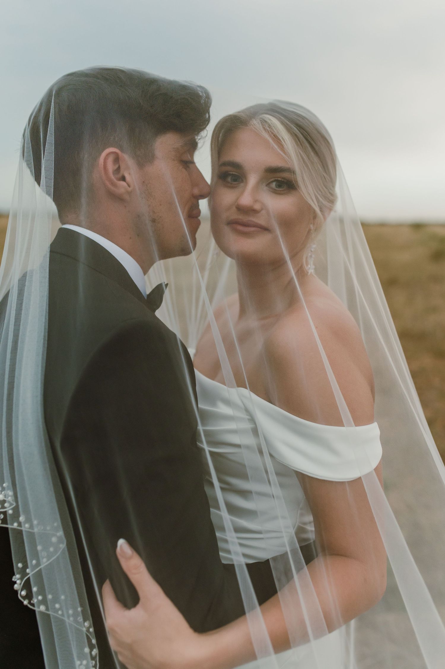Elegant wedding couple poses with delicate veil in afternoon light.