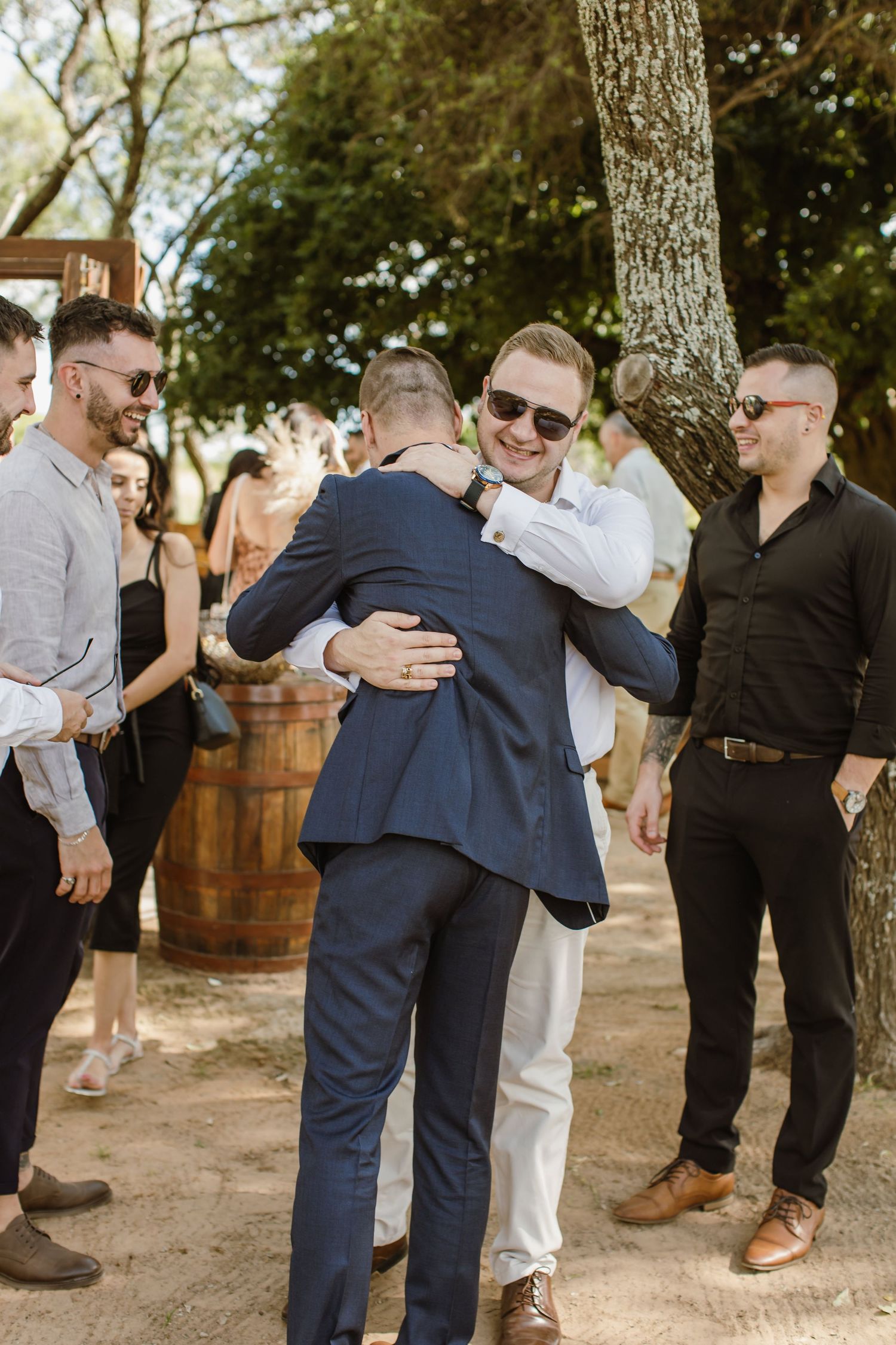 Wedding celebration with guests hugging and embracing outdoors under trees.