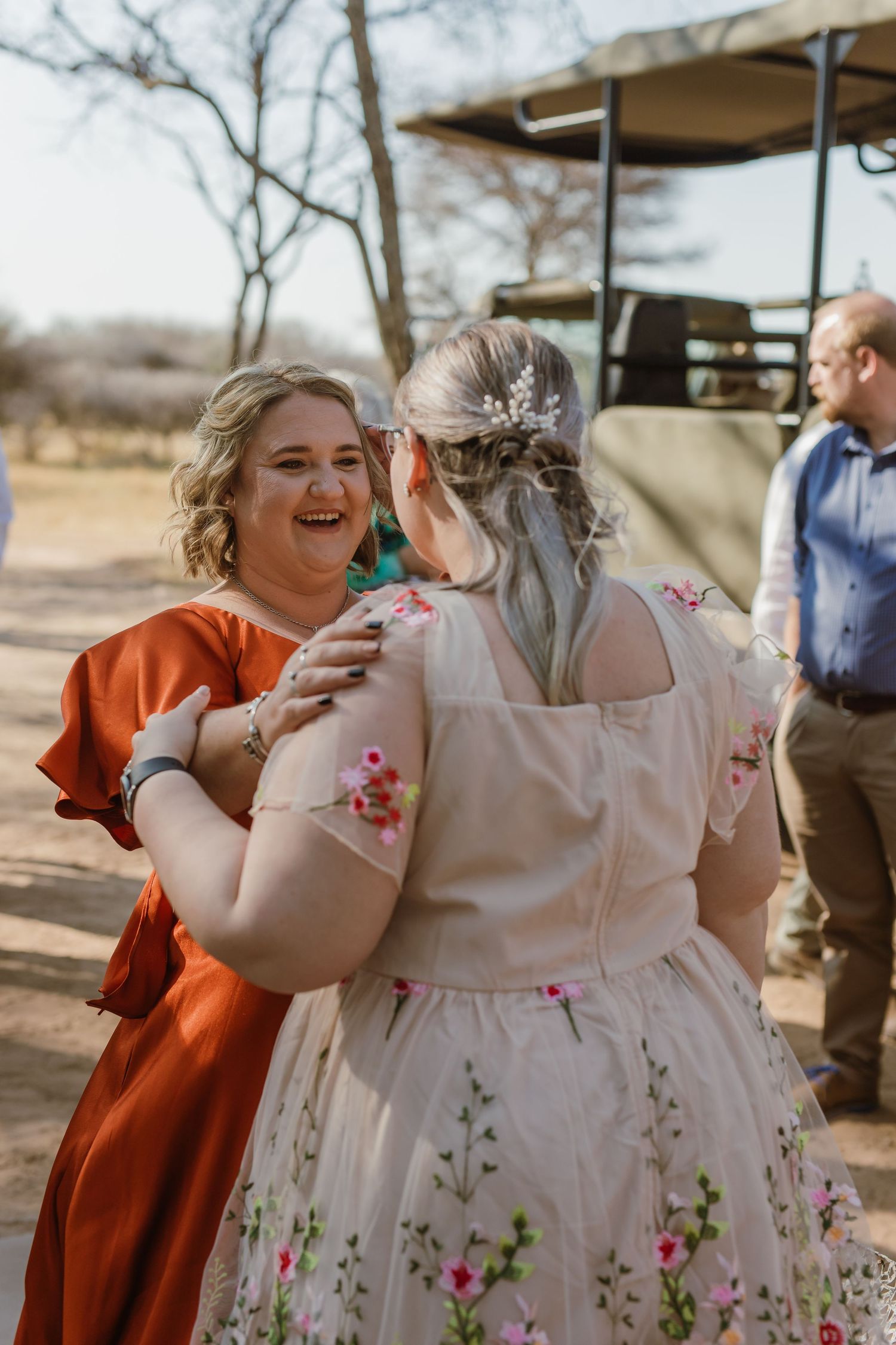 Wedding guests in colorful dresses embrace and laugh during an outdoor celebration.
