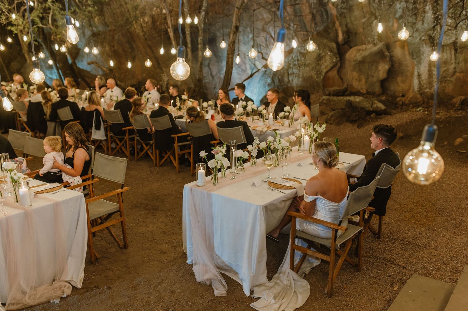 Outdoor wedding reception setup with white tables and string lights hanging between trees.