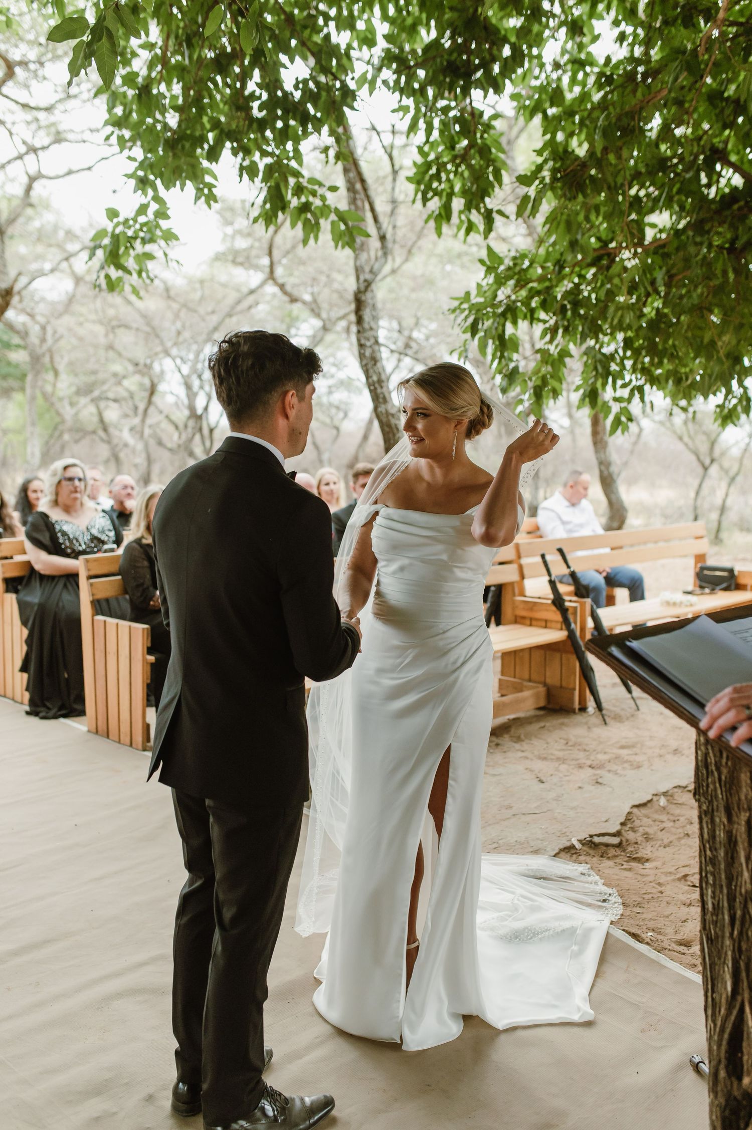 Outdoor wedding ceremony with wooden bench seating and natural decor under tree coverage.