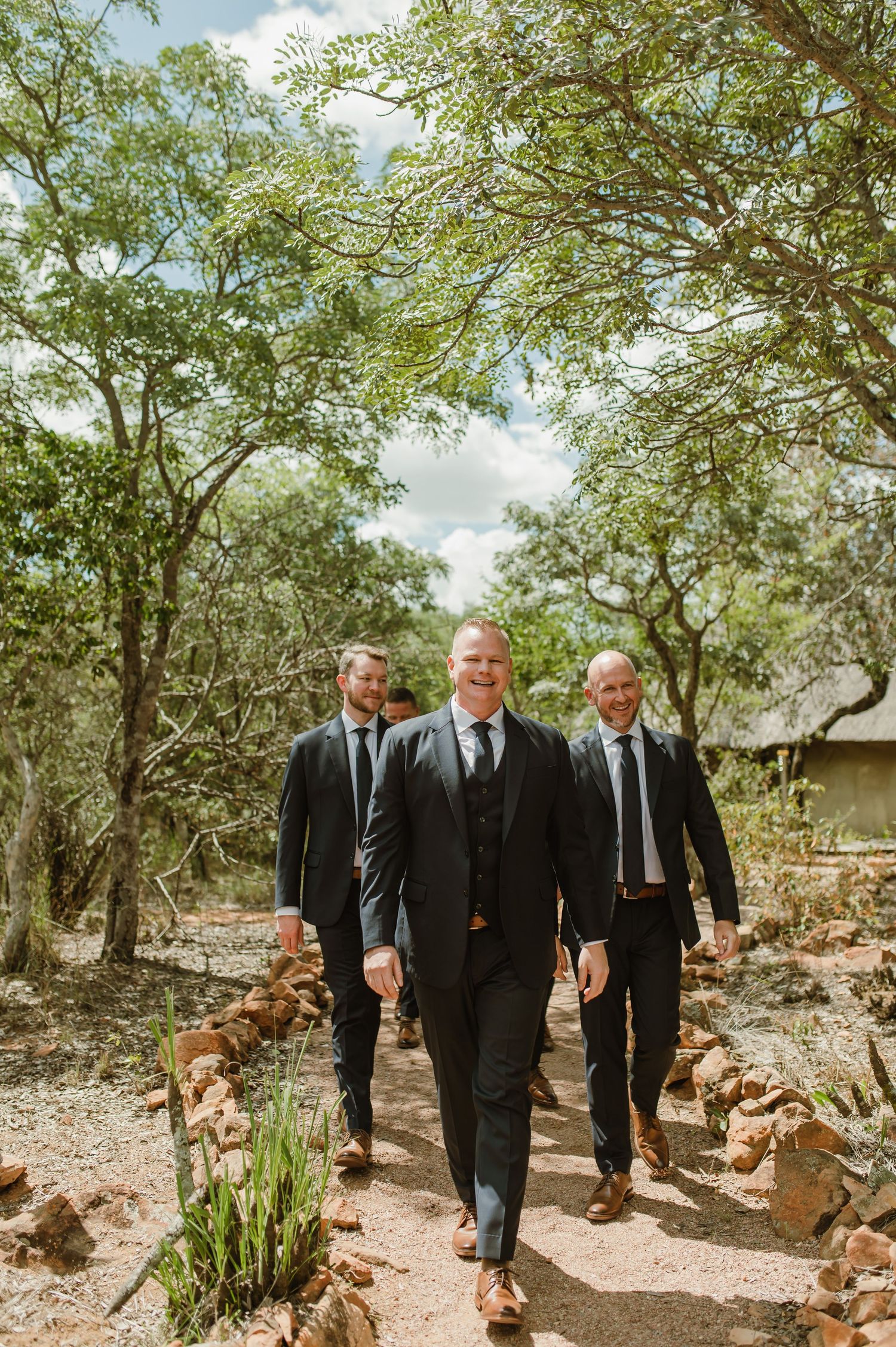 Groomsmen walking together through a sunlit garden path in formal black suits.