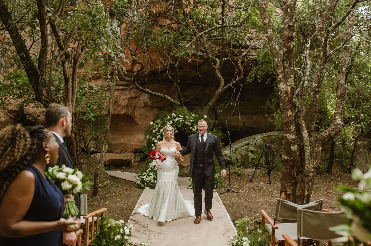 Natural outdoor wedding venue with wooden chairs arranged under towering rock formations.