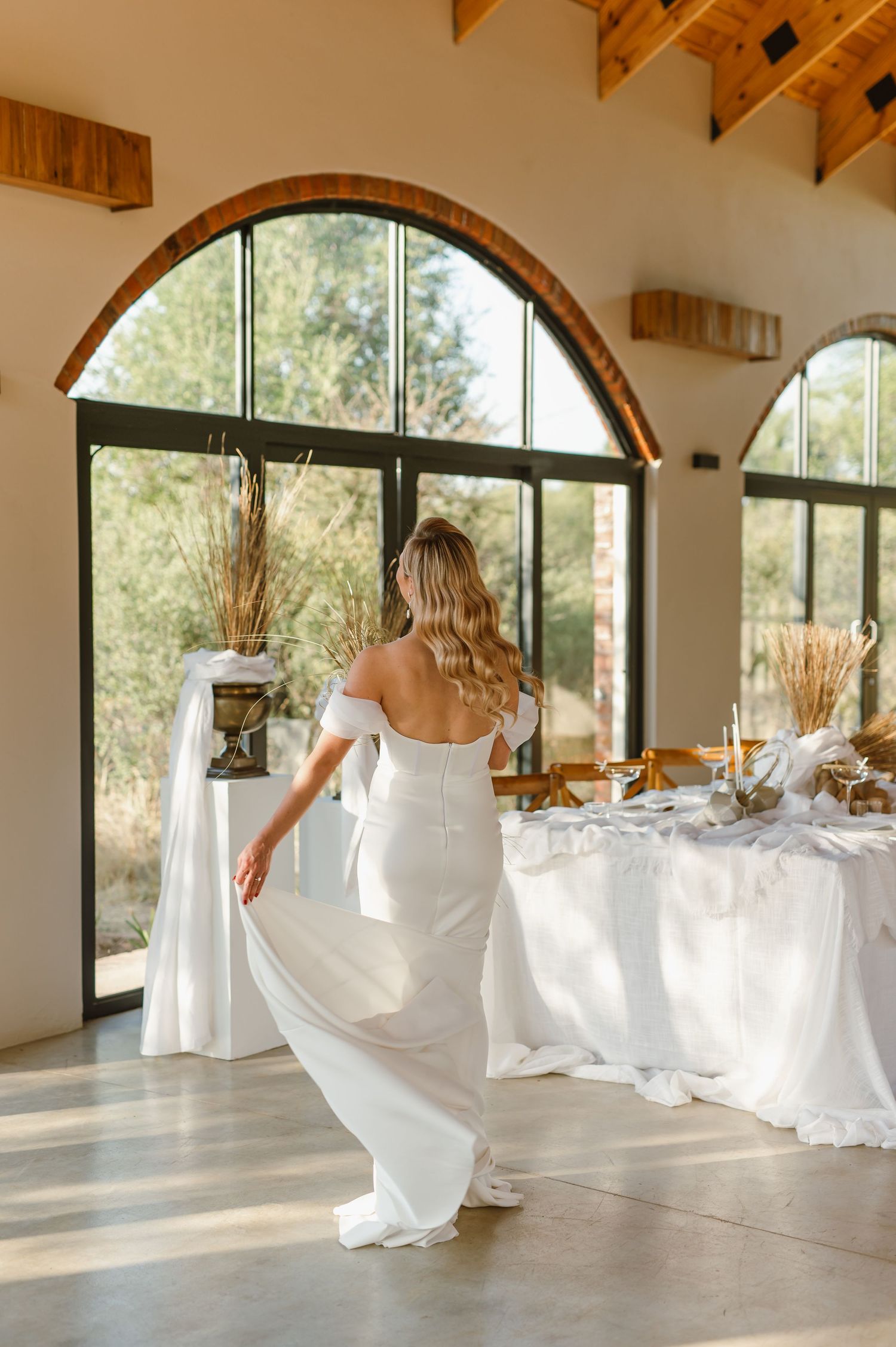 Elegant white wedding reception setup with flowing tablecloths in a bright venue with arched windows.