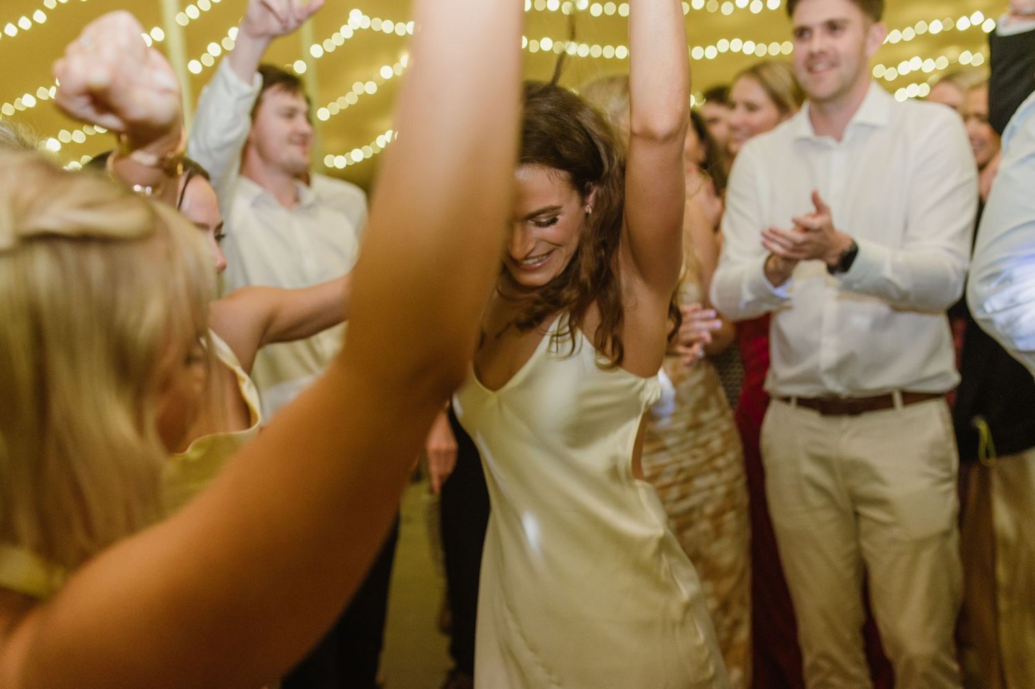 Energetic dance floor scene with bridesmaids in light colored dresses under string lights.