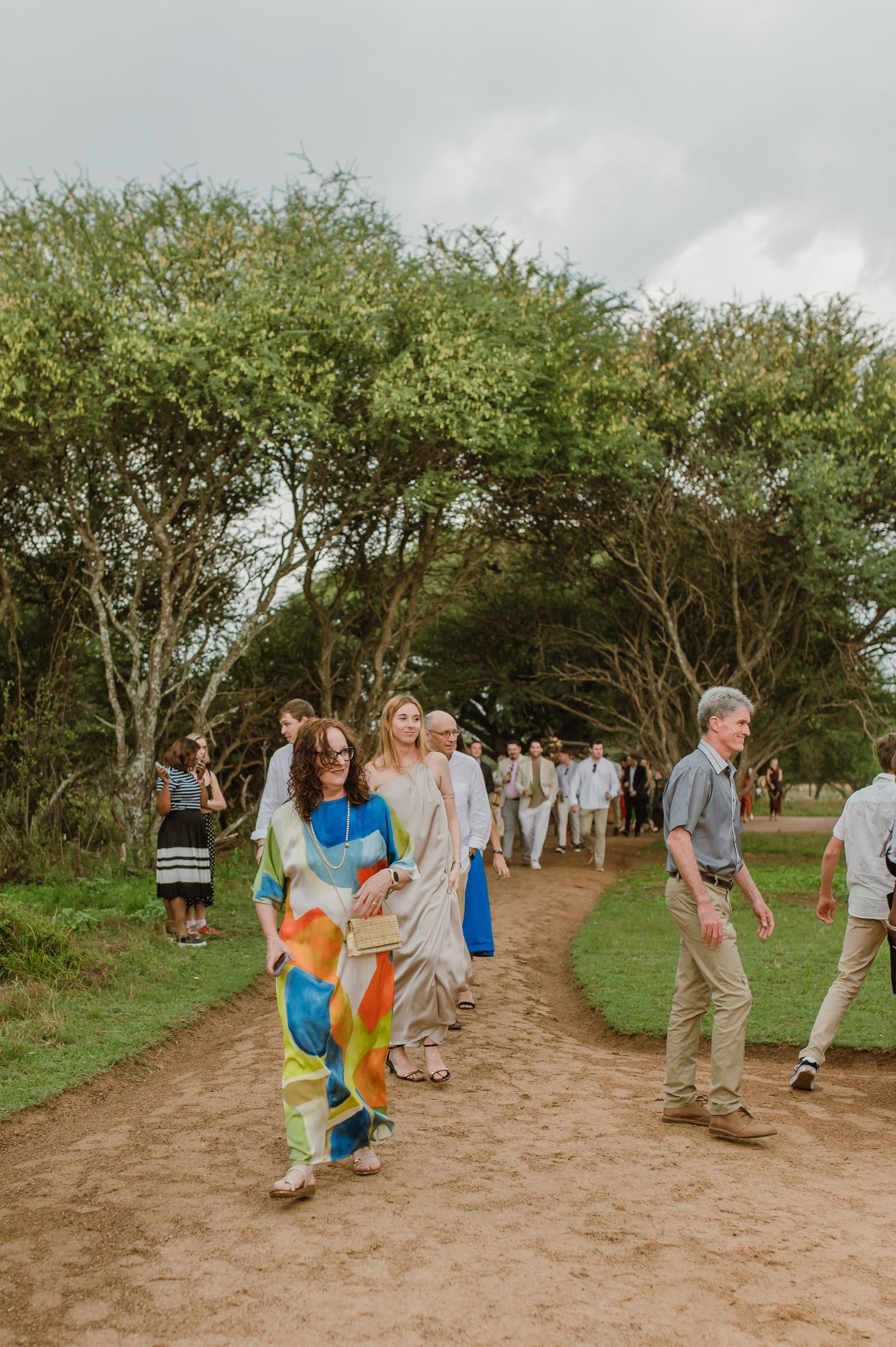 Wedding attendees walking along a dirt path lined with trees and greenery.