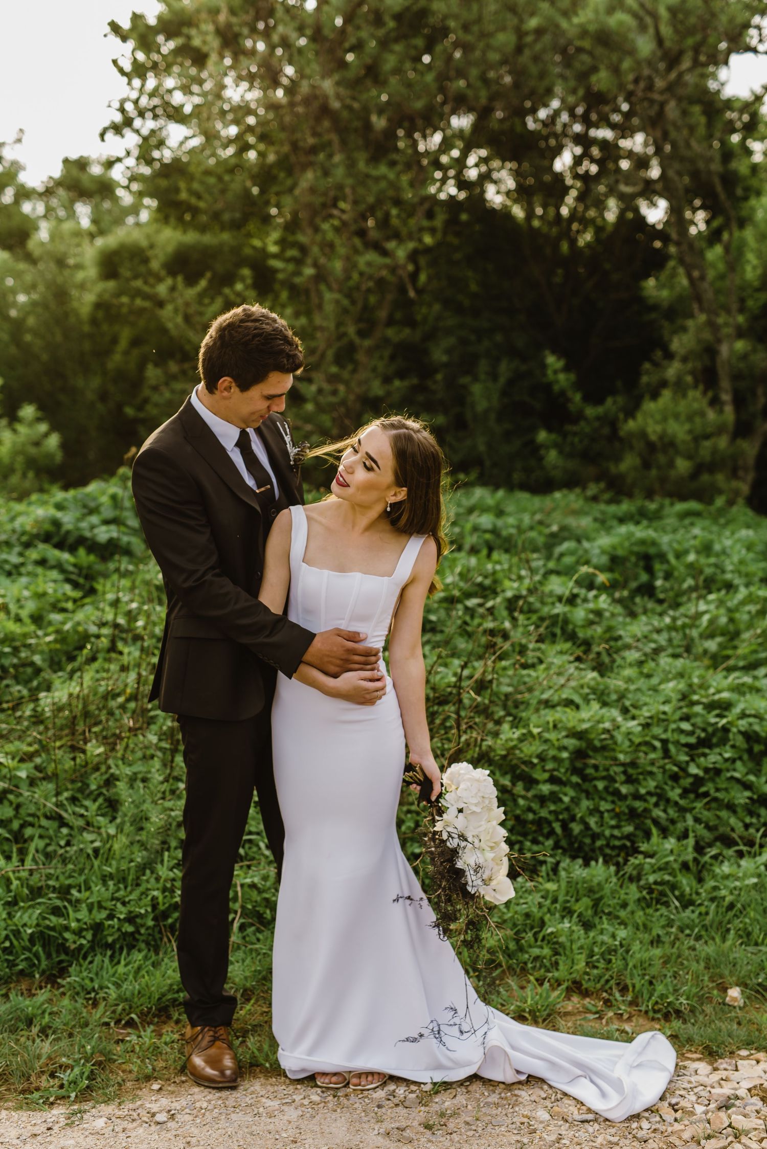 A couple shares an intimate moment on a rustic path, with bride in white dress and groom in black suit.