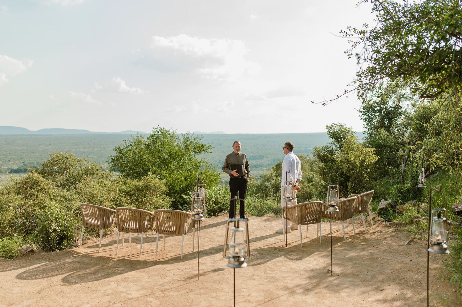 Two people standing at an outdoor wedding ceremony venue overlooking a scenic African savanna landscape.