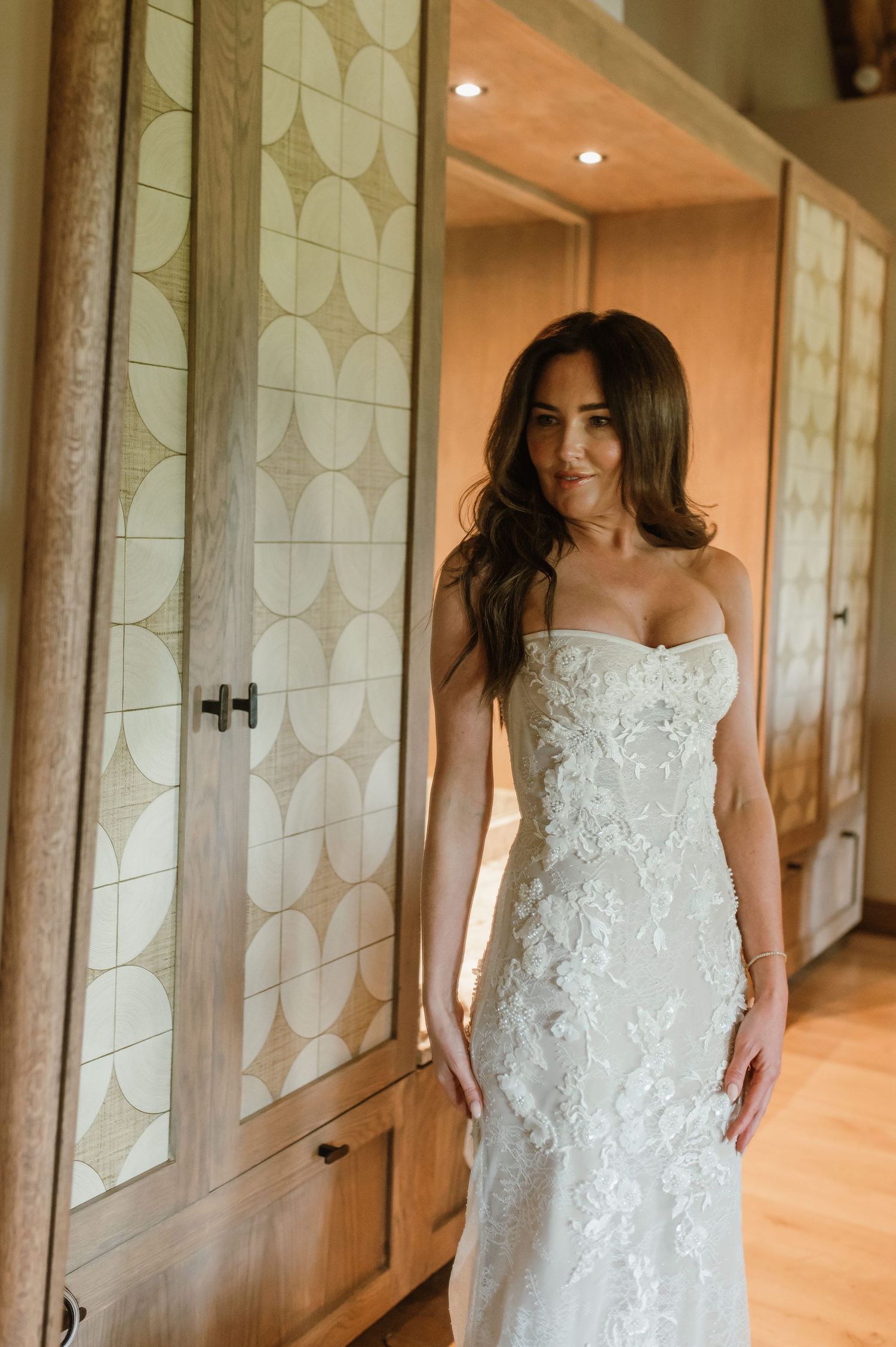 Beautiful bride in a strapless floral lace wedding gown posing beside decorative wardrobe doors.